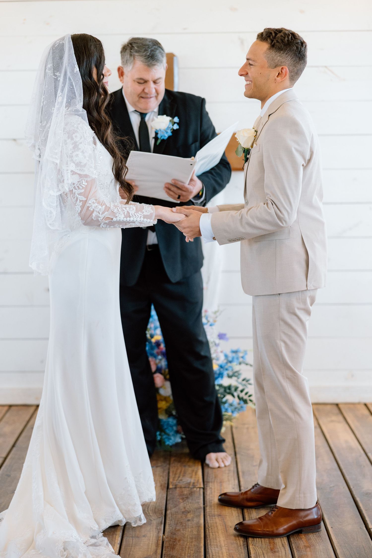 Bride and groom exchange vows holding hands before officiant at rustic white chapel wedding ceremony.