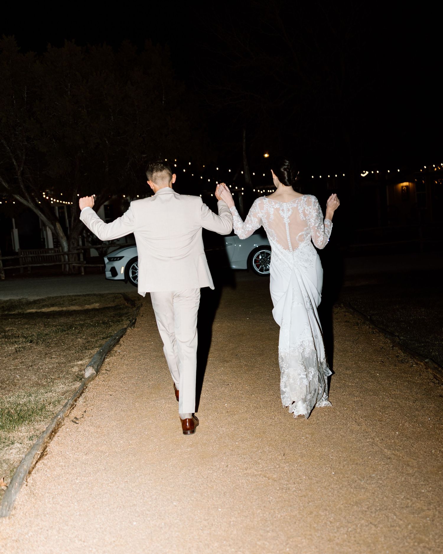 Bride and groom walking hand in hand down a path at night with string lights overhead.