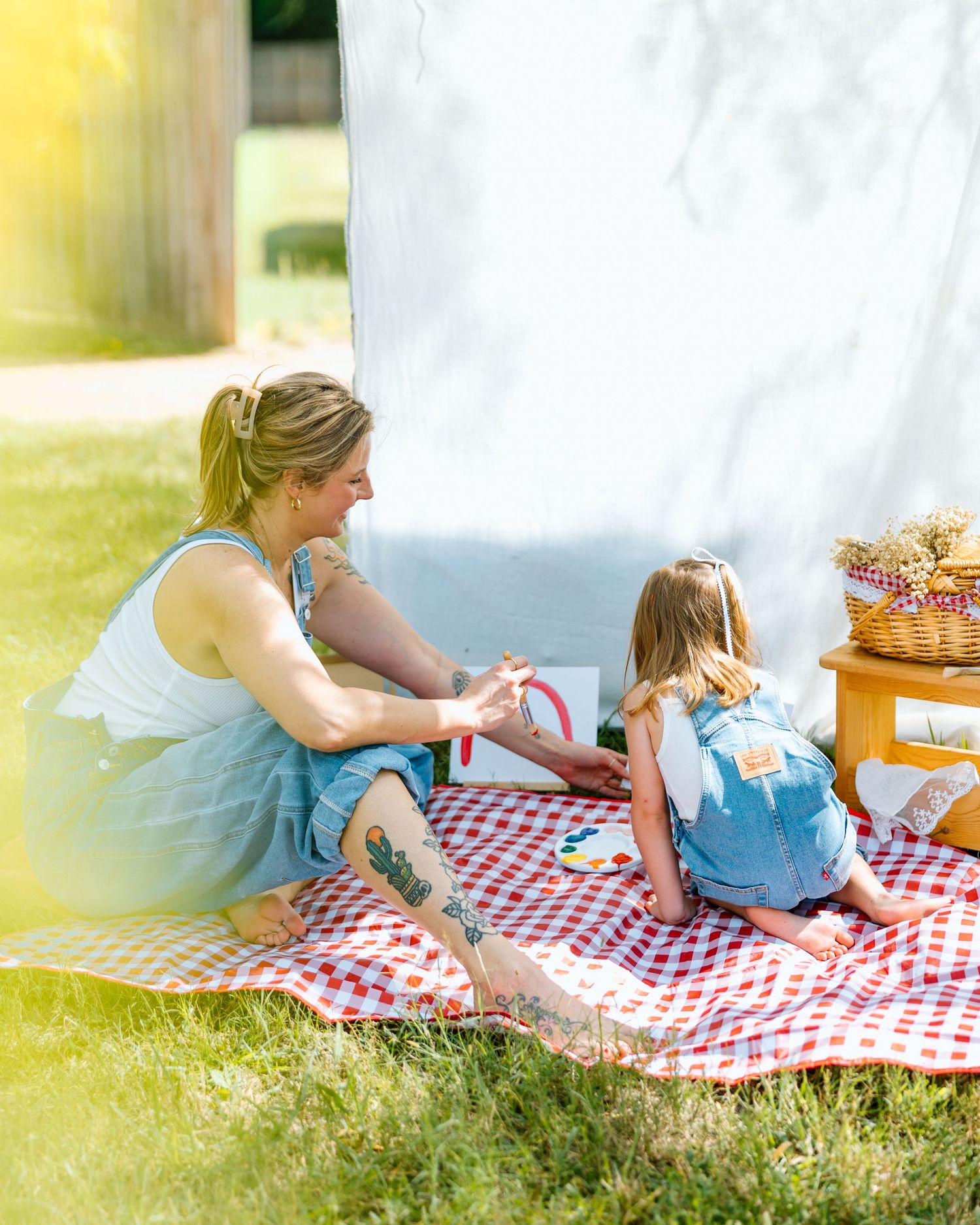 Mom and daughter in denim overalls sit on red gingham picnic blanket outdoors, playing together on sunny day.