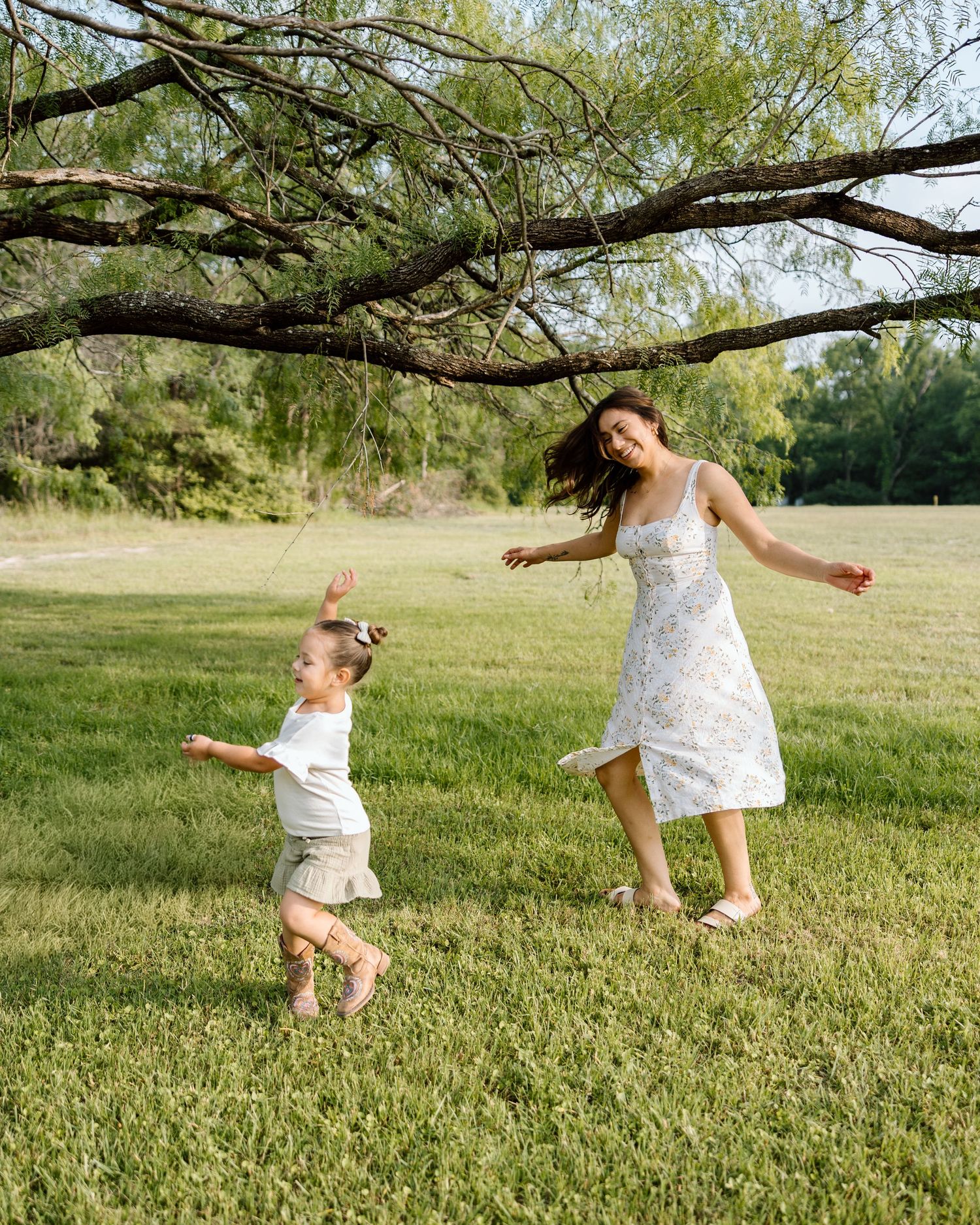 A woman in a floral dress plays with a toddler on a green lawn beneath a tree on a sunny day.