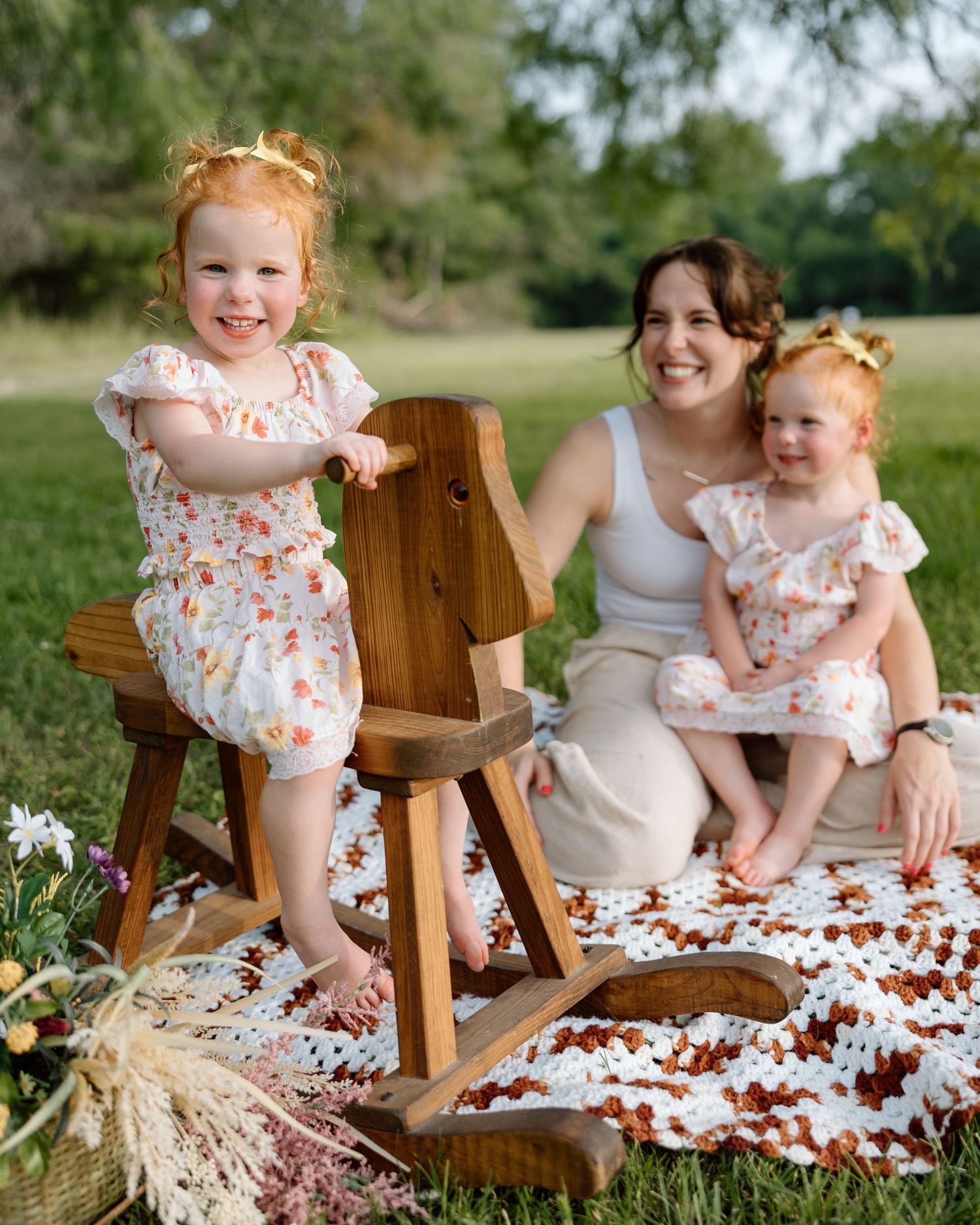 A mother sits on a crochet blanket outdoors with two young redhead girls in matching floral outfits near a wooden rocking horse.