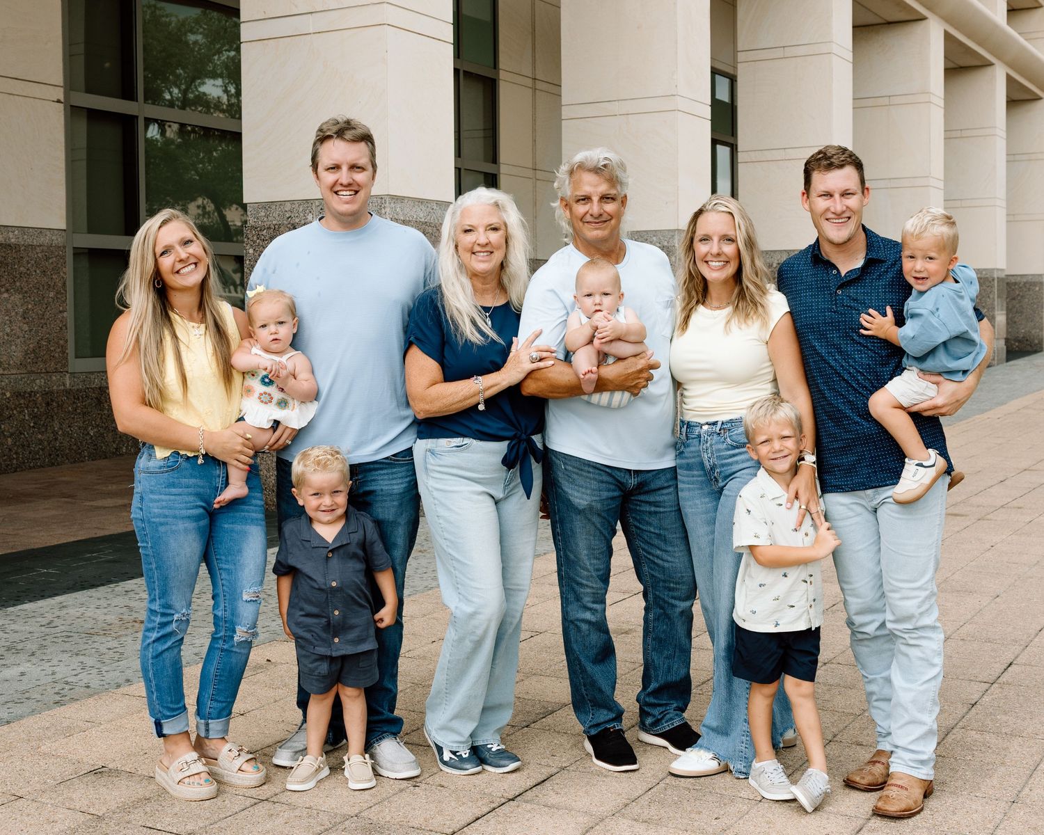 Multi-generational family portrait with grandparents, parents, and young children smiling outdoors near a building.