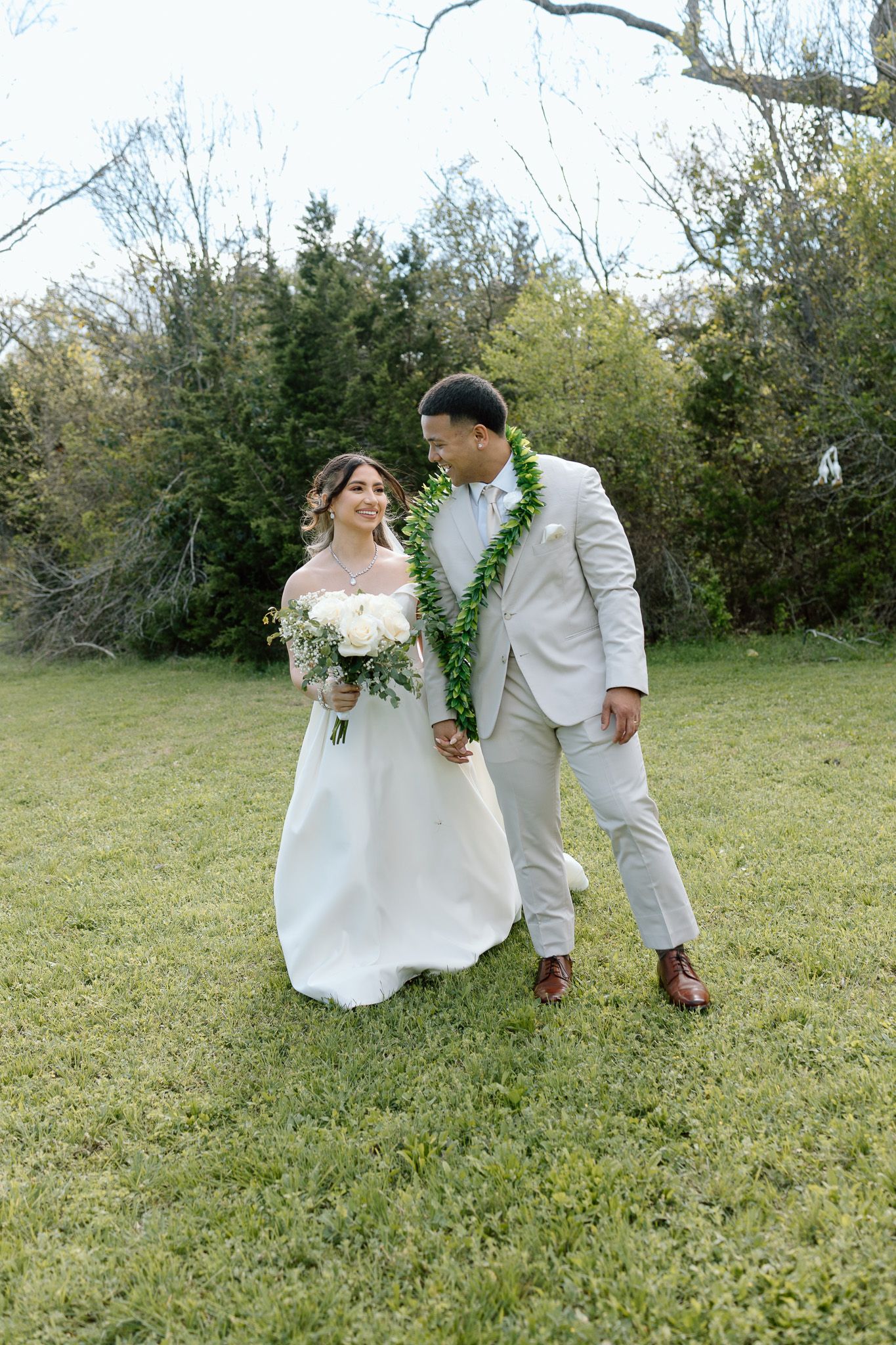 Newlyweds in white attire and green leis walk together on grass during their outdoor wedding.