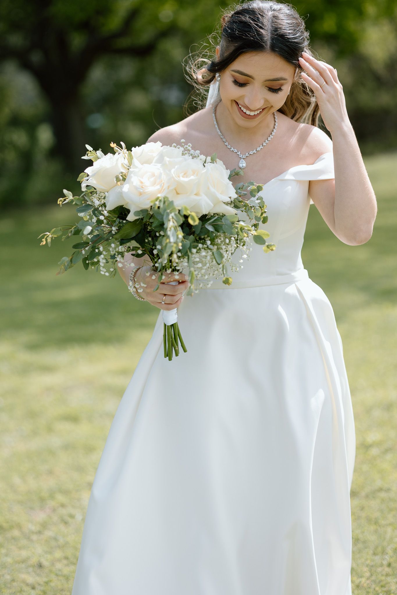 Bride in off-shoulder gown holds white bouquet while smiling in sunlit garden setting.