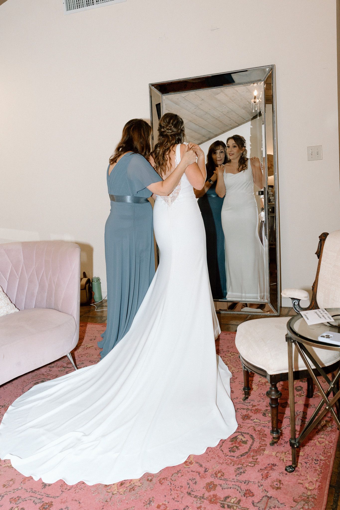 Bridesmaids help with a bride's wedding dress train while looking in a large mirror.