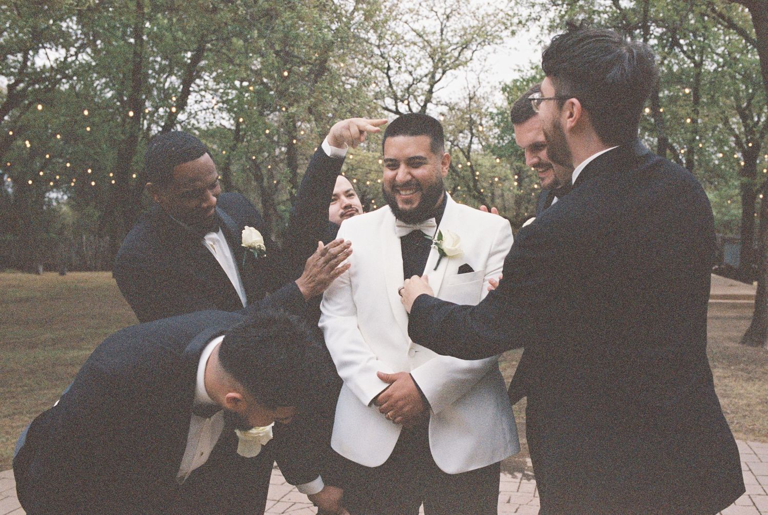 Groomsmen in suits playfully lift and celebrate on an outdoor brick pathway.