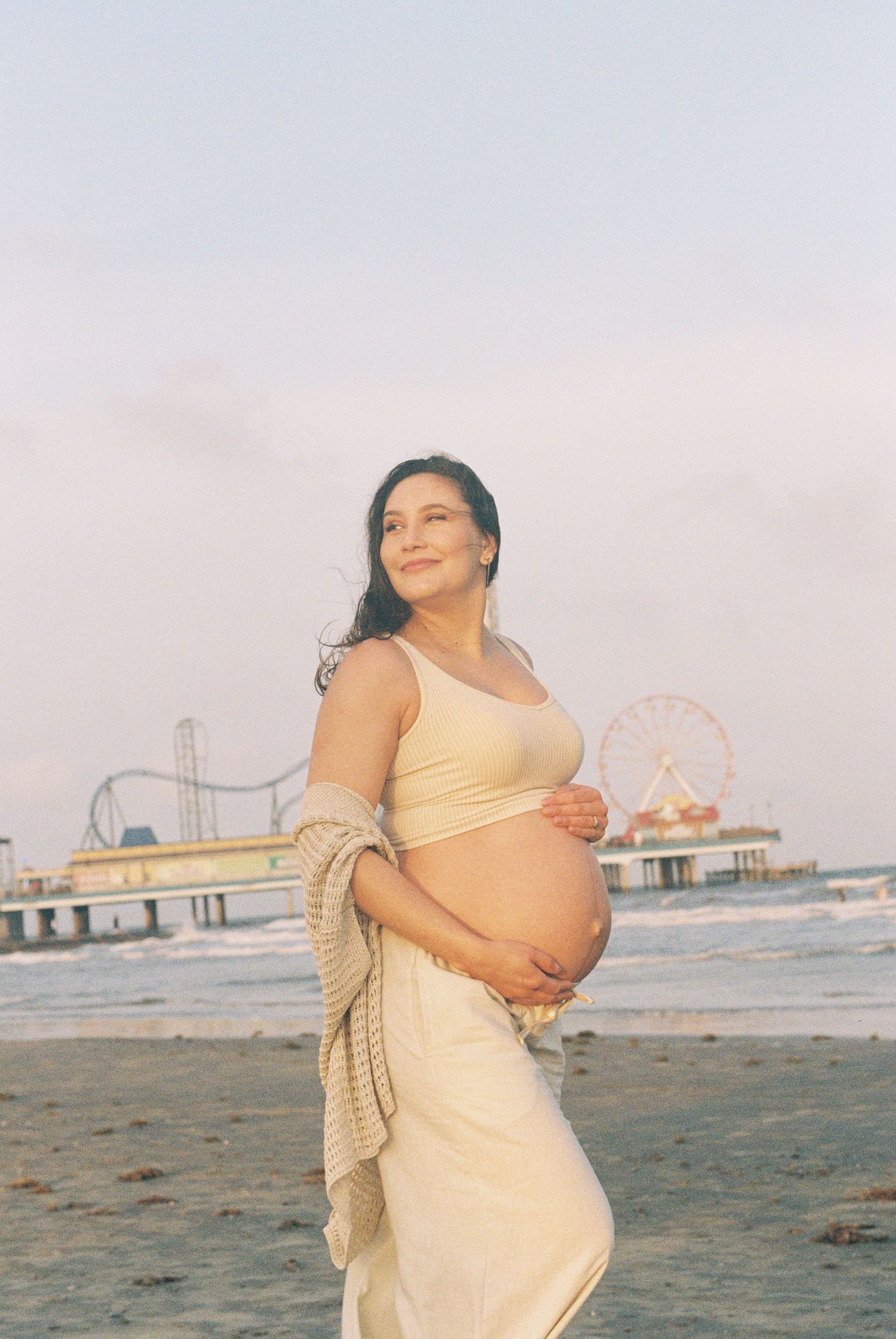 Expectant mother in yellow dress poses on beach with amusement park pier and ferris wheel in background.