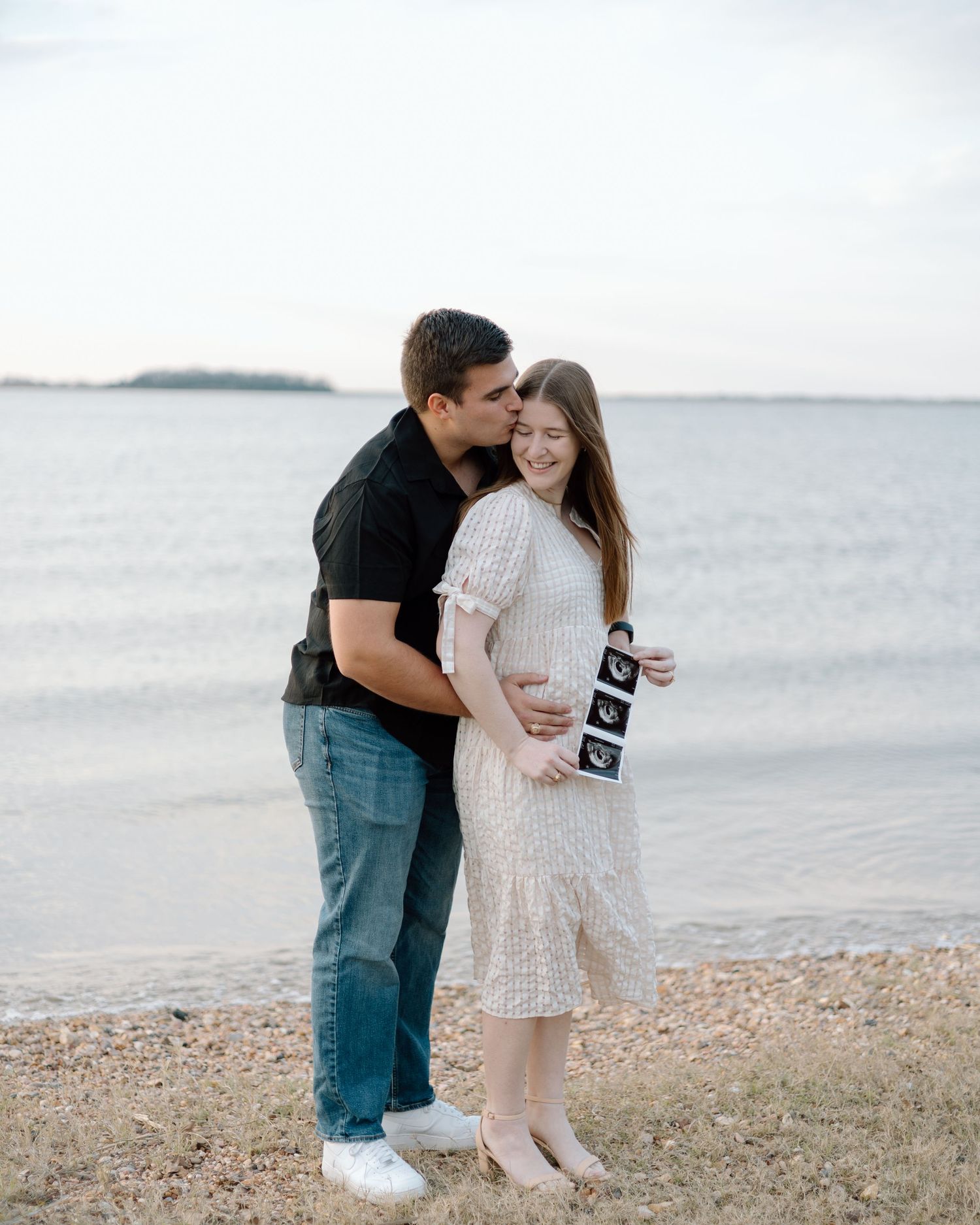A couple embraces on a coastal shoreline while holding ultrasound images.