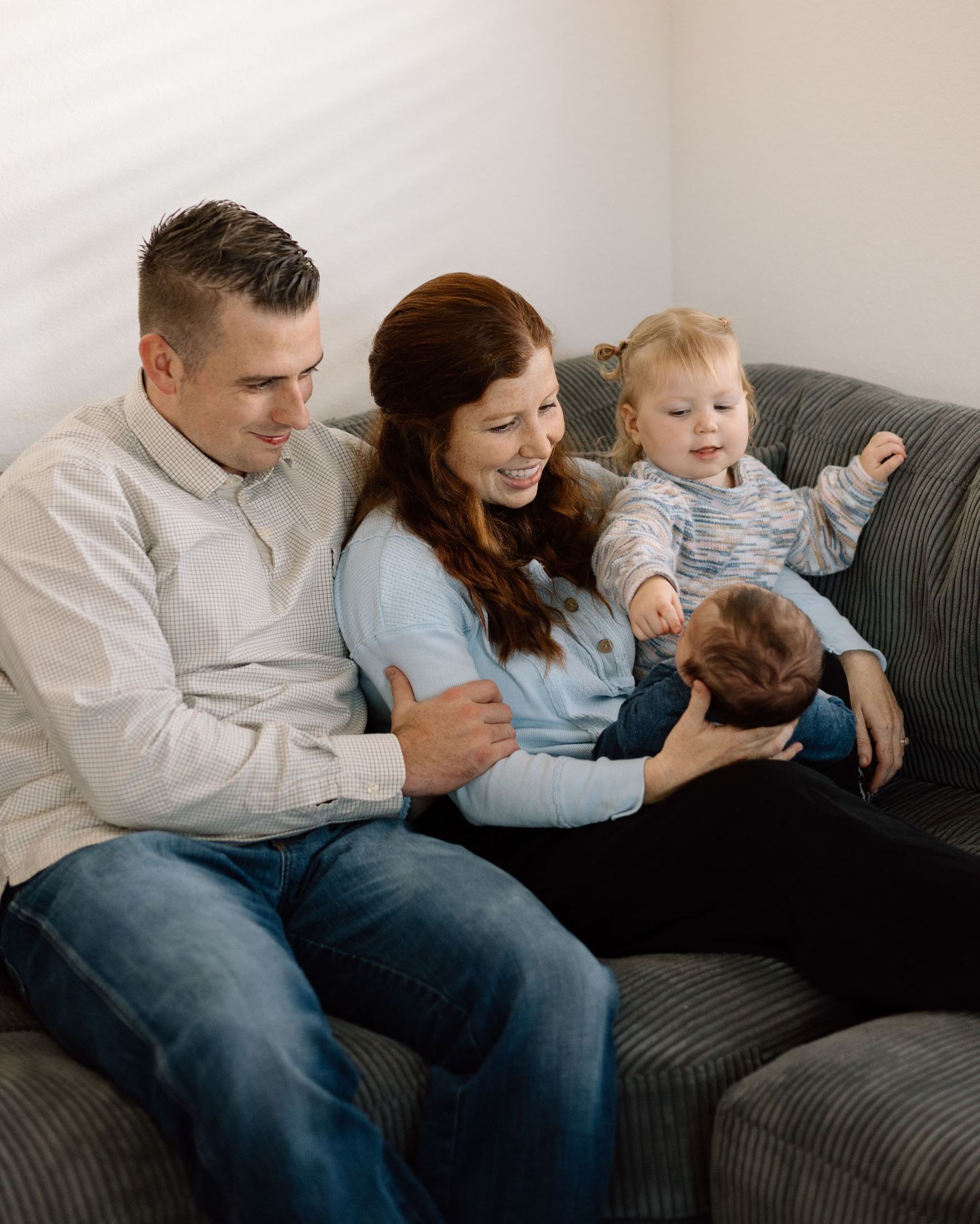 Happy family of four sitting on a gray couch, parents smiling at newborn baby while toddler plays beside them.