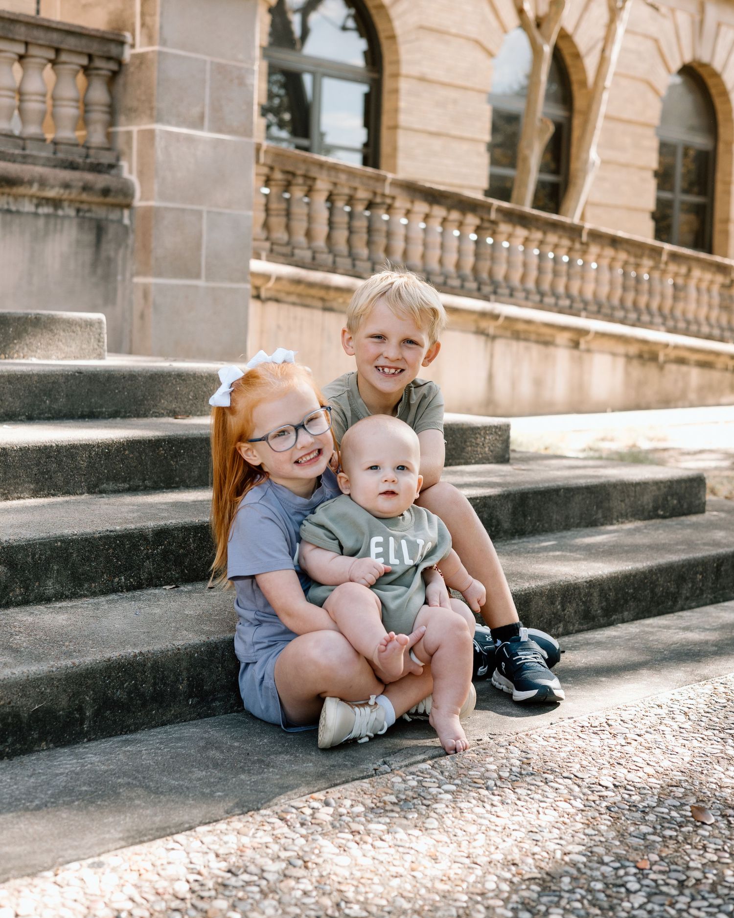 Three young siblings sitting on stone steps, two older children holding a baby, smiling outdoors near a historic building.