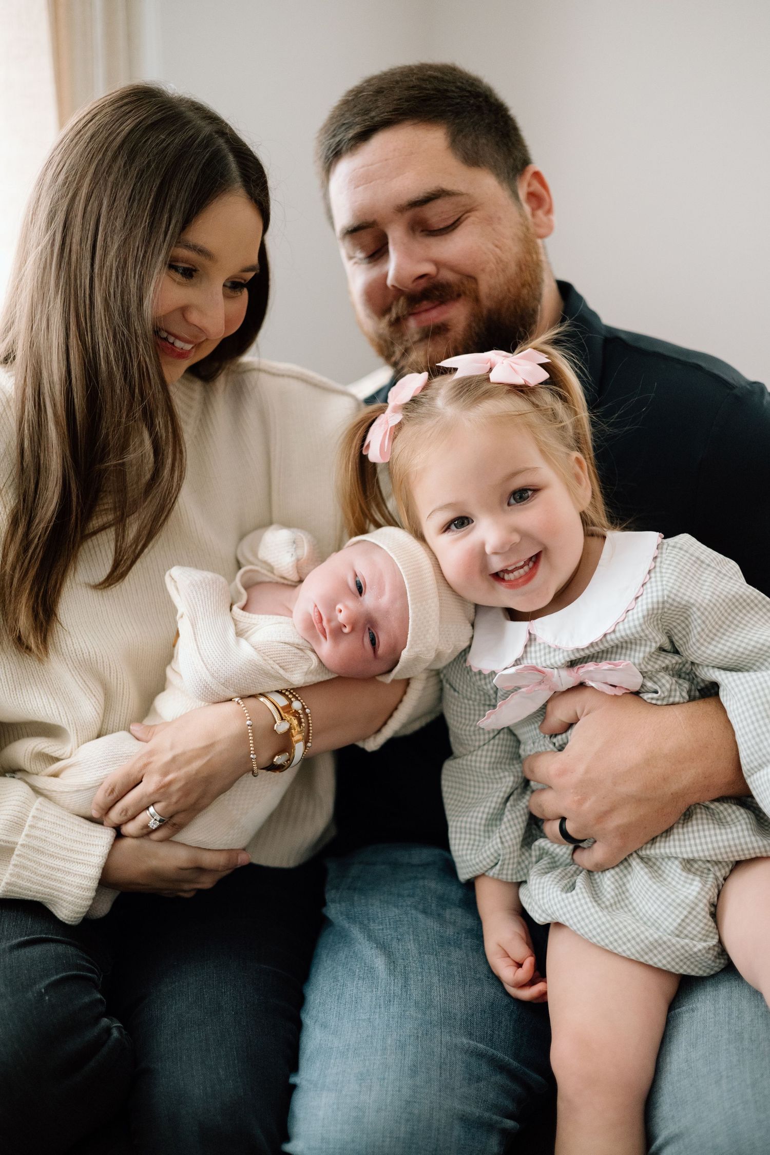 Happy family of four with mom holding newborn baby and dad embracing smiling toddler girl with pink bows.