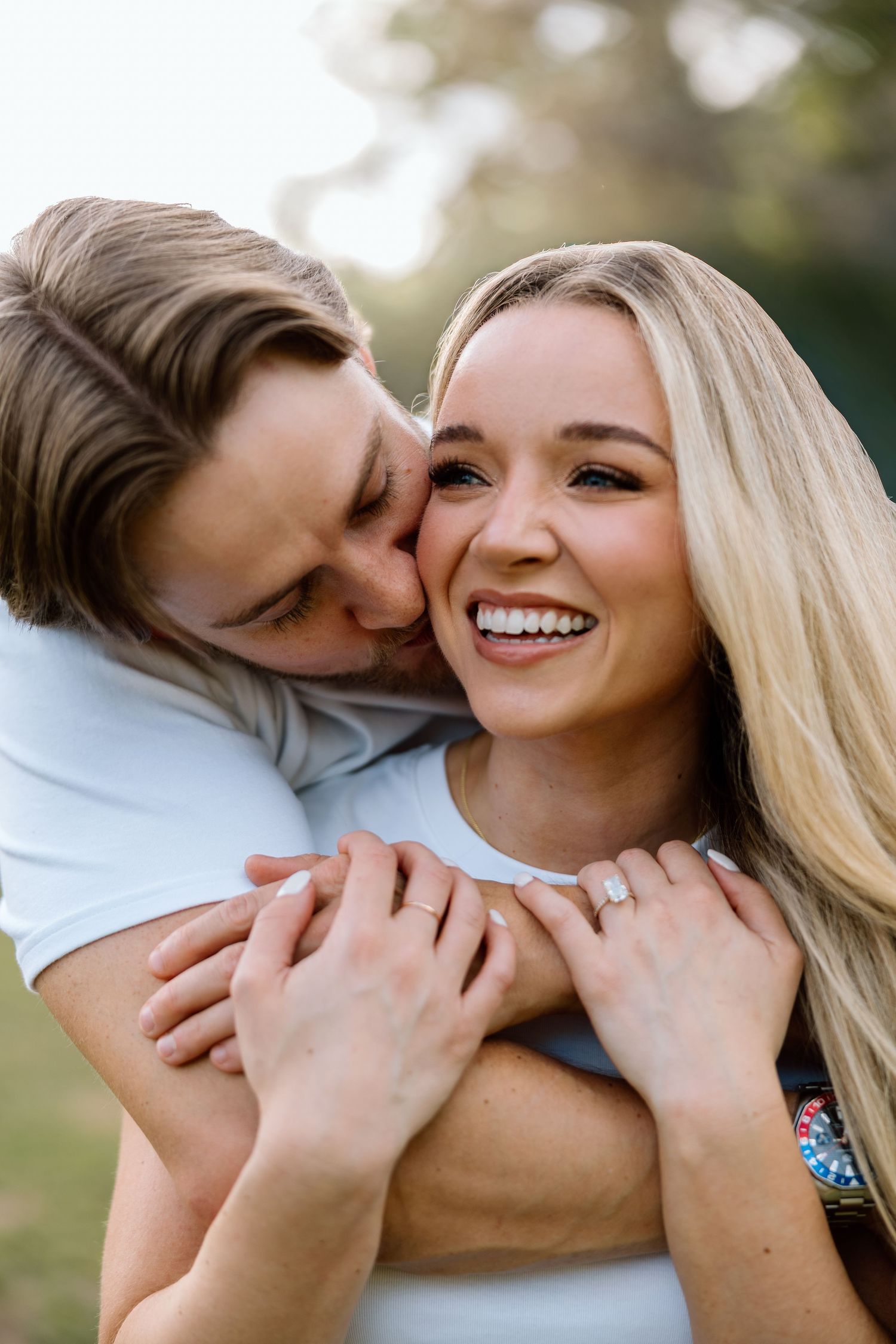 Couple sharing a joyful moment as man kisses woman's cheek while embracing her from behind outdoors.