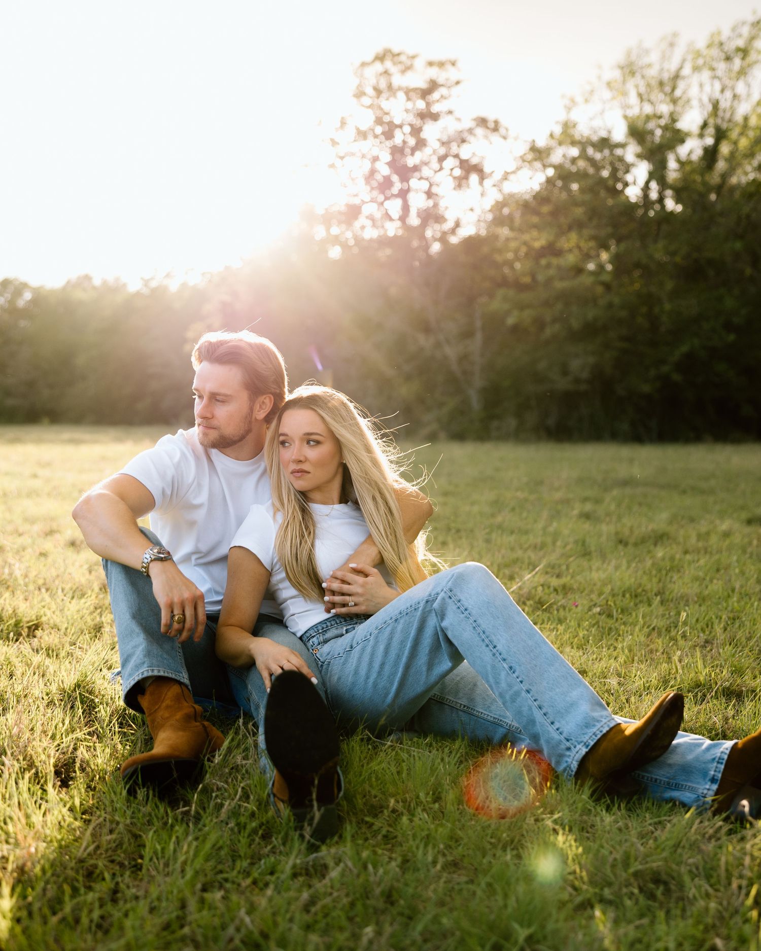 Couple sitting together in a golden sunlit field, wearing white tees and light jeans, enjoying a peaceful moment.