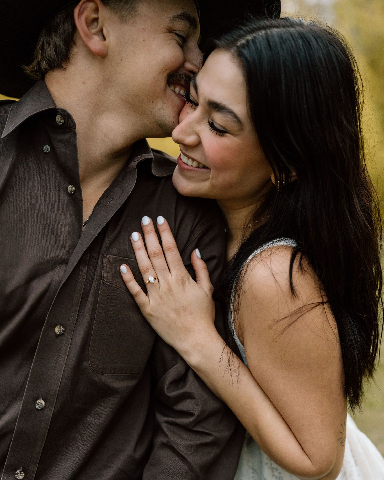 Happy couple sharing a romantic moment, woman in white dress showing engagement ring, man in brown shirt and cowboy hat.