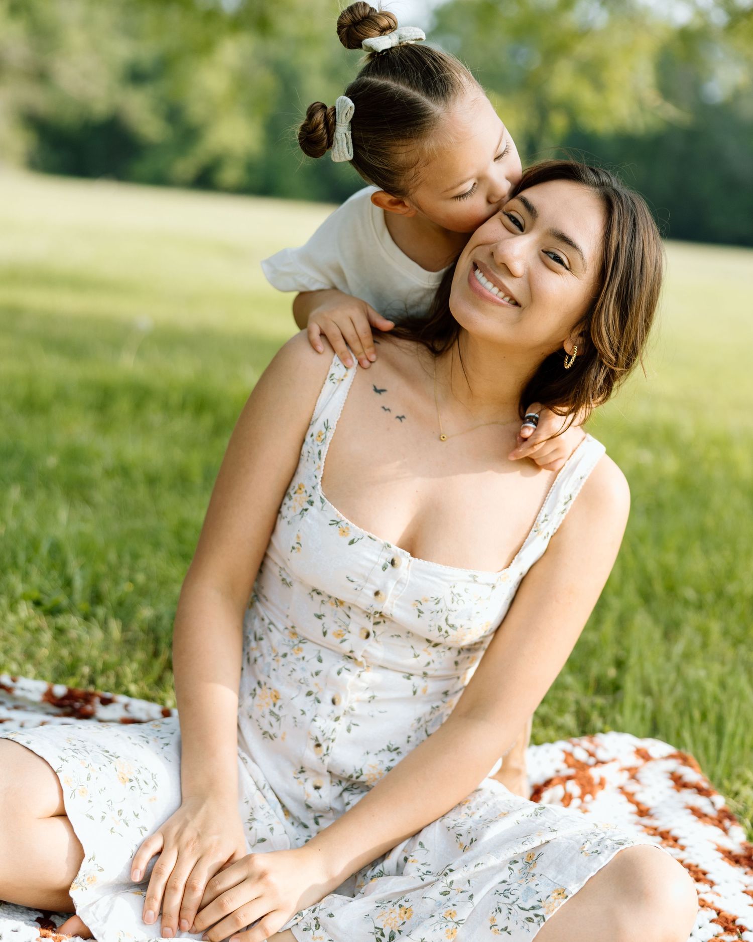 A young woman in a floral dress sits on a blanket outdoors as a toddler girl hugs and kisses her cheek.
