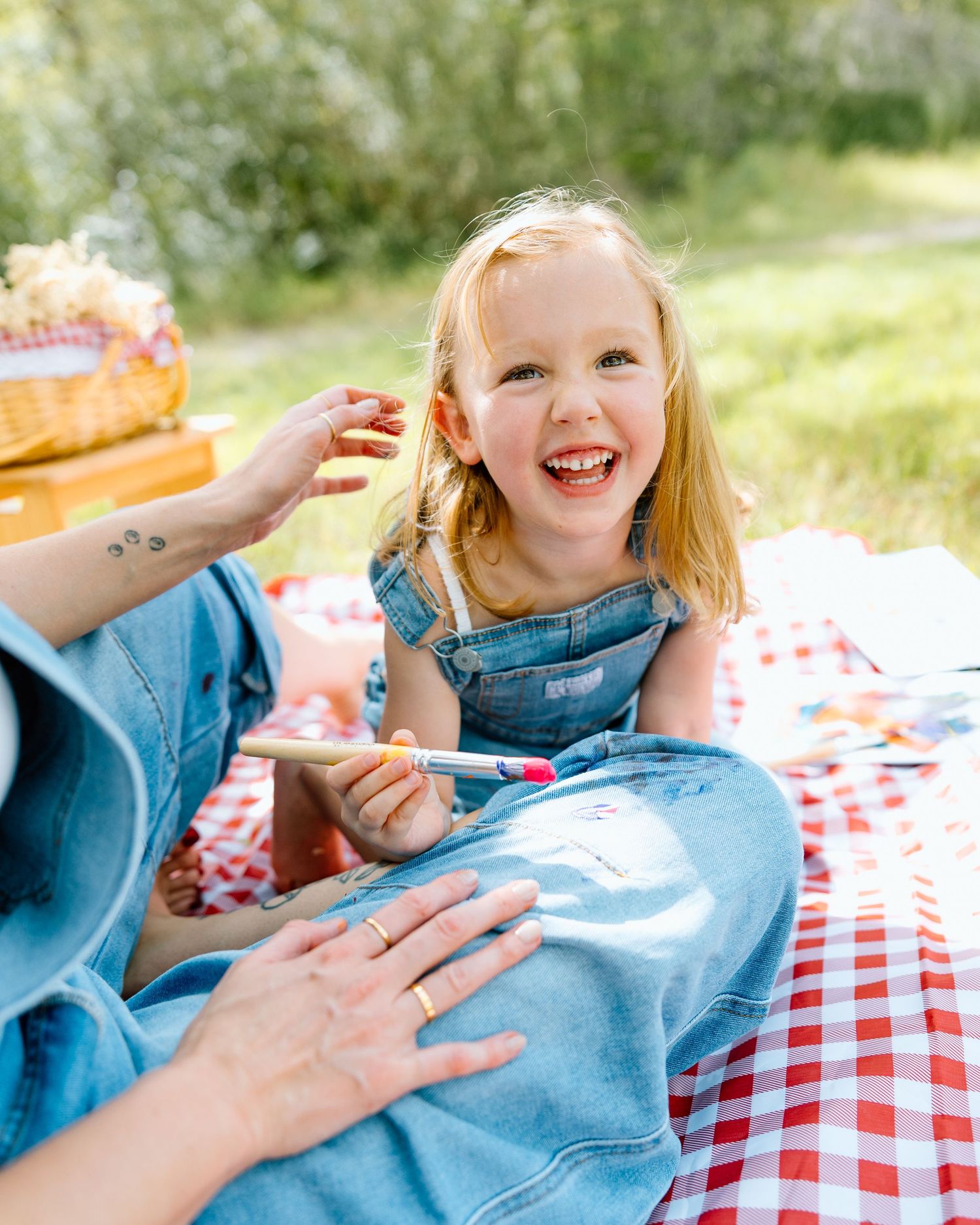 A happy young girl in denim overalls laughs during a sunny outdoor picnic on a red checkered blanket.
