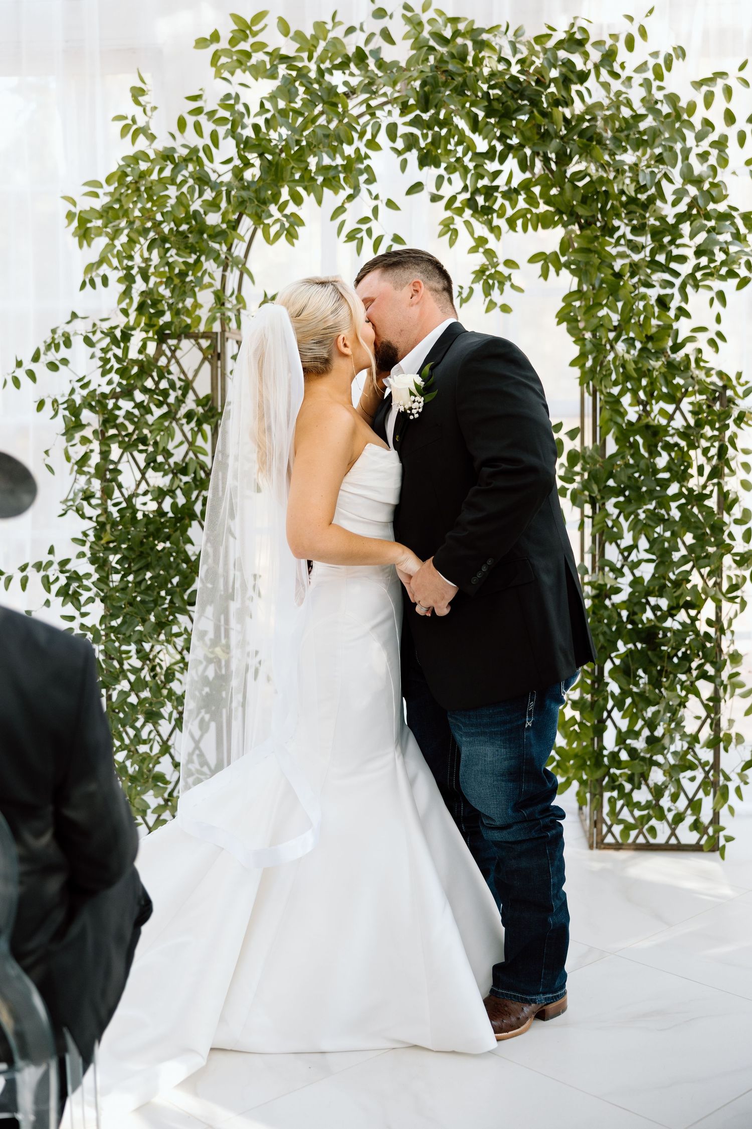 Bride and groom share first kiss under green leaf arch at outdoor wedding ceremony.