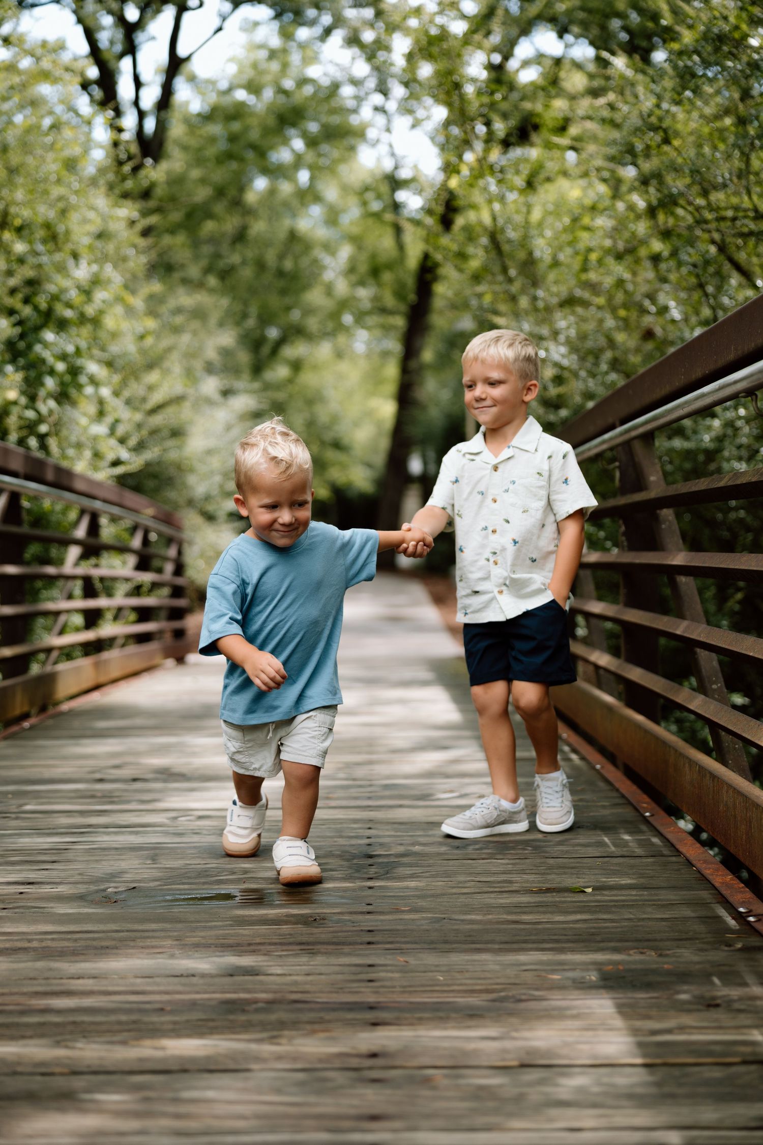 Two young blonde boys holding hands and walking on a wooden bridge surrounded by lush green trees.