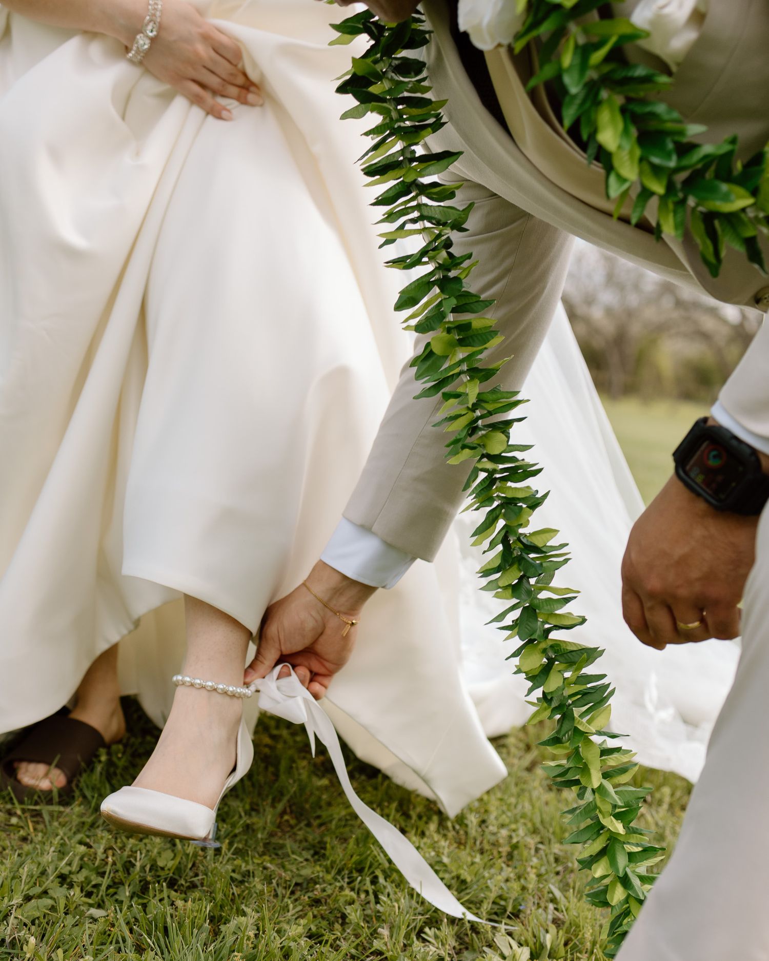 Detail shot of wedding attire showing green lei garland and bride's white shoes being adjusted.