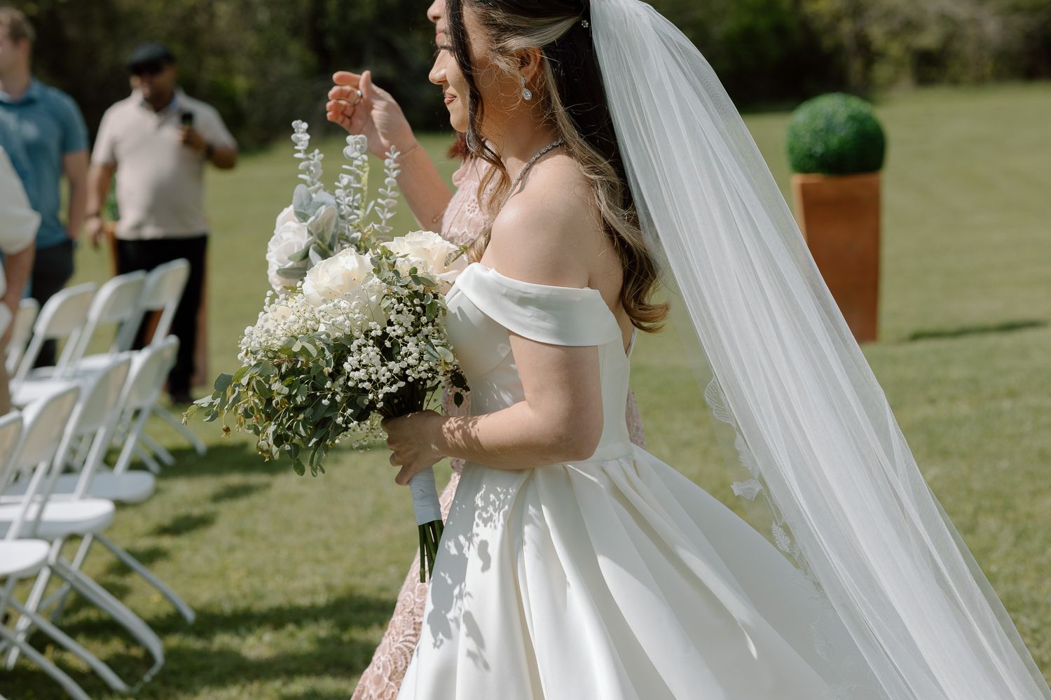 Bride in an elegant off-shoulder wedding dress holds a white bouquet during an outdoor garden ceremony.