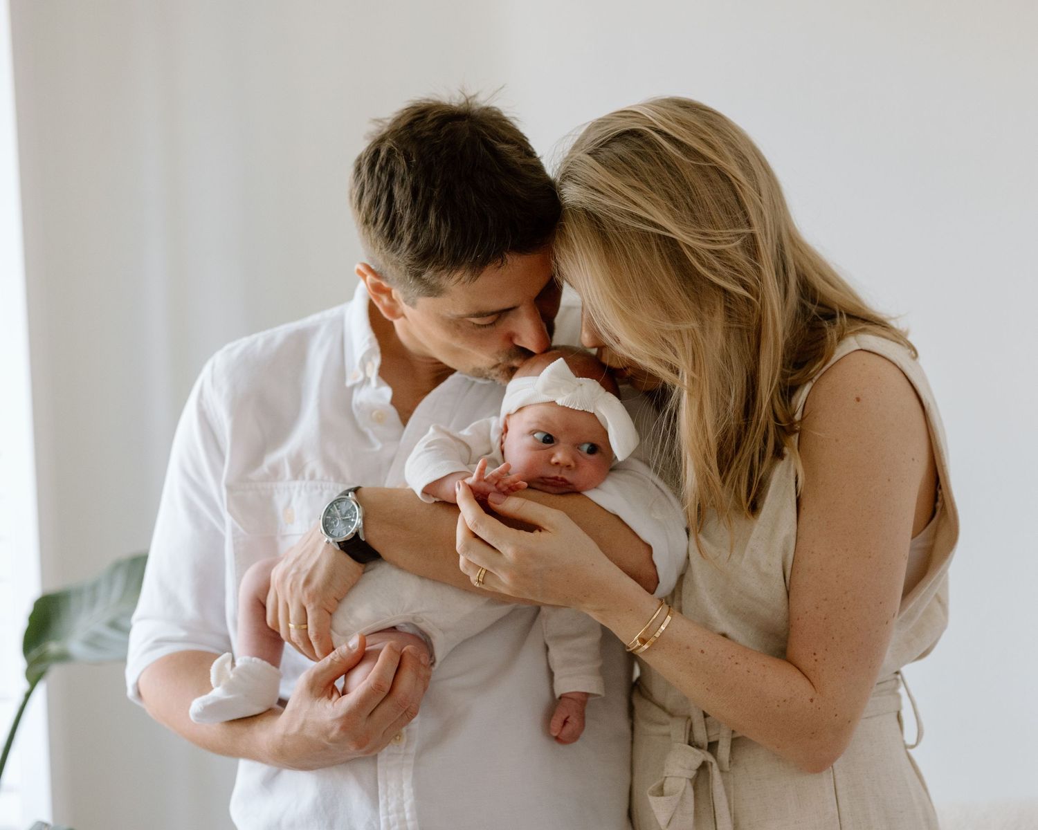 New parents in white outfits share intimate moment while holding their newborn.
