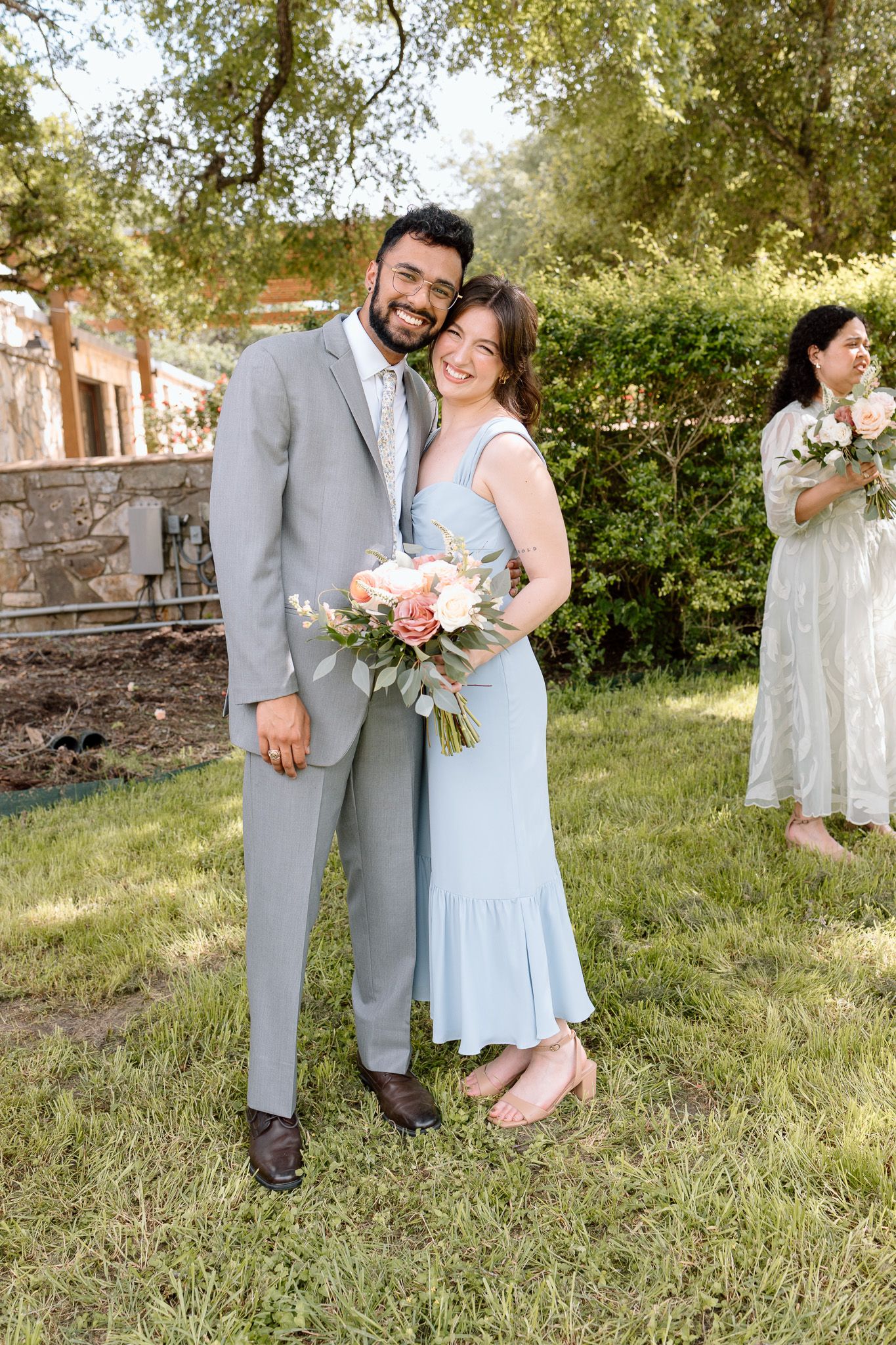 A couple in light blue and gray attire pose together at an outdoor wedding venue with greenery in the background.