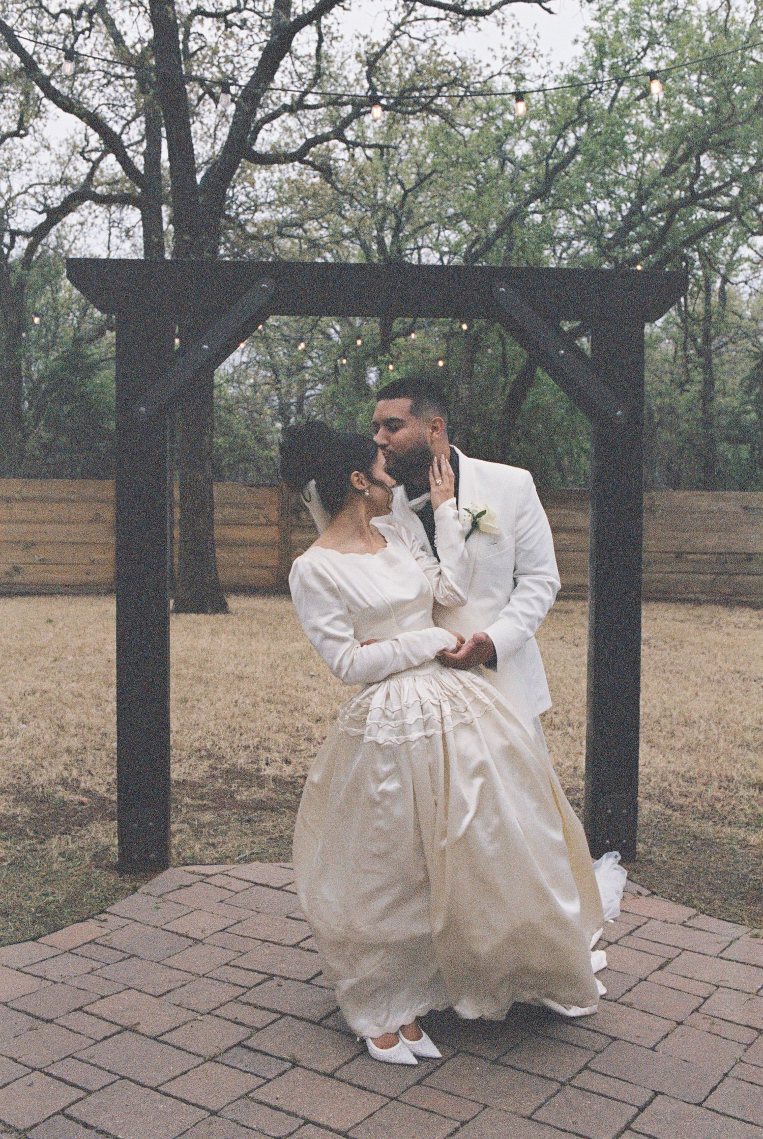 A romantic wedding couple embraces under a wooden pergola in a natural outdoor setting.