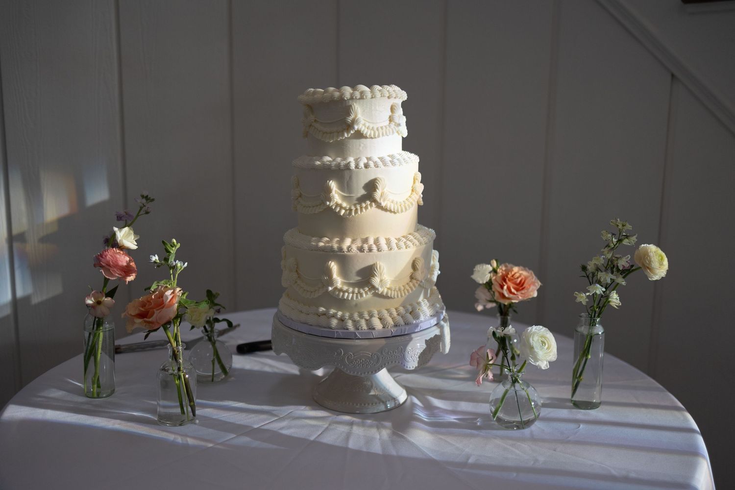 White wedding cake displayed on table with floral arrangements in bud vases.