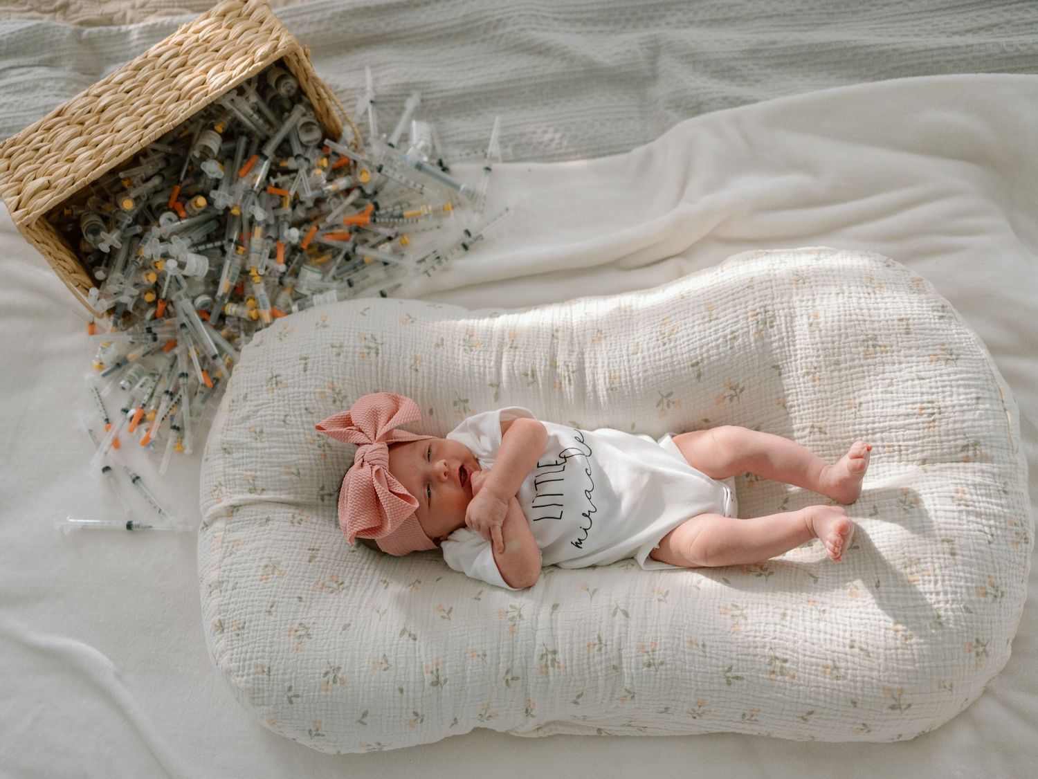 Ballet pointe shoes and pink fabric bow rest on a delicate white linen surface beside a woven basket with dried flowers.