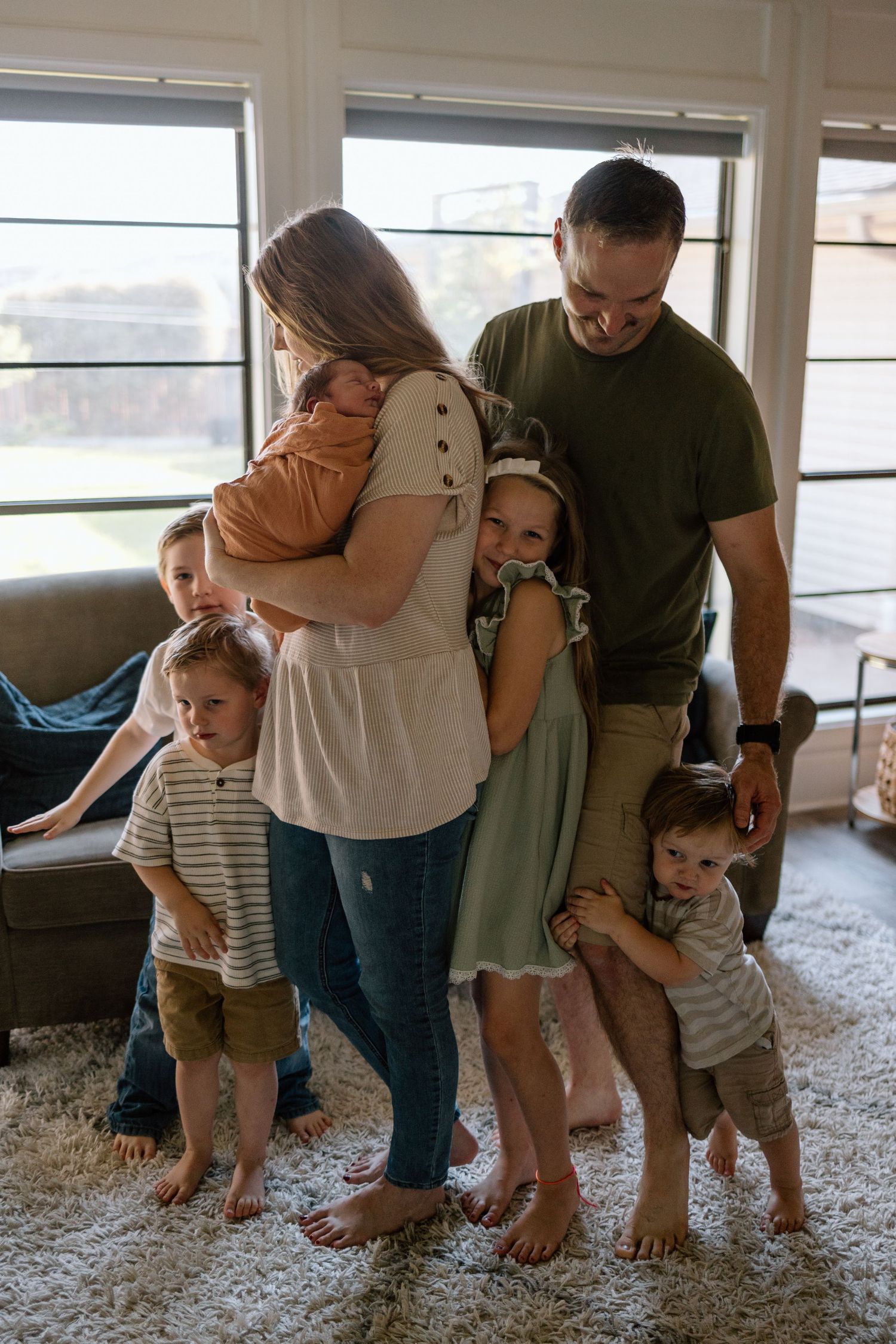 A family stands together in a cozy living room with carpeted floors and large windows, sharing an intimate moment.