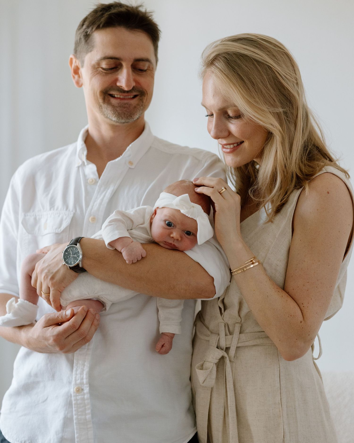 New parents in white and beige outfits holding their newborn baby with joy.