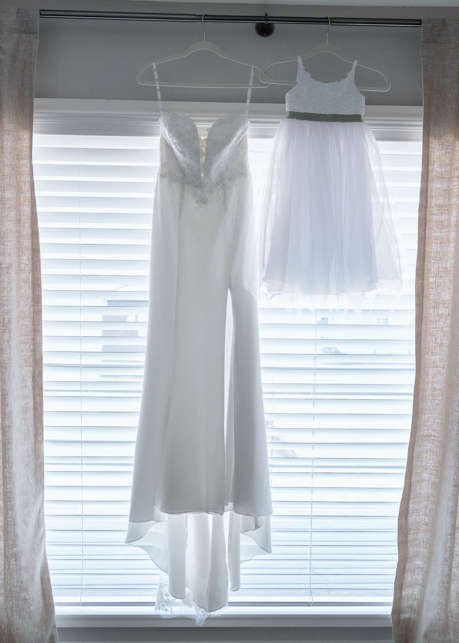 White wedding dress and jumpsuit hanging by window with venetian blinds.