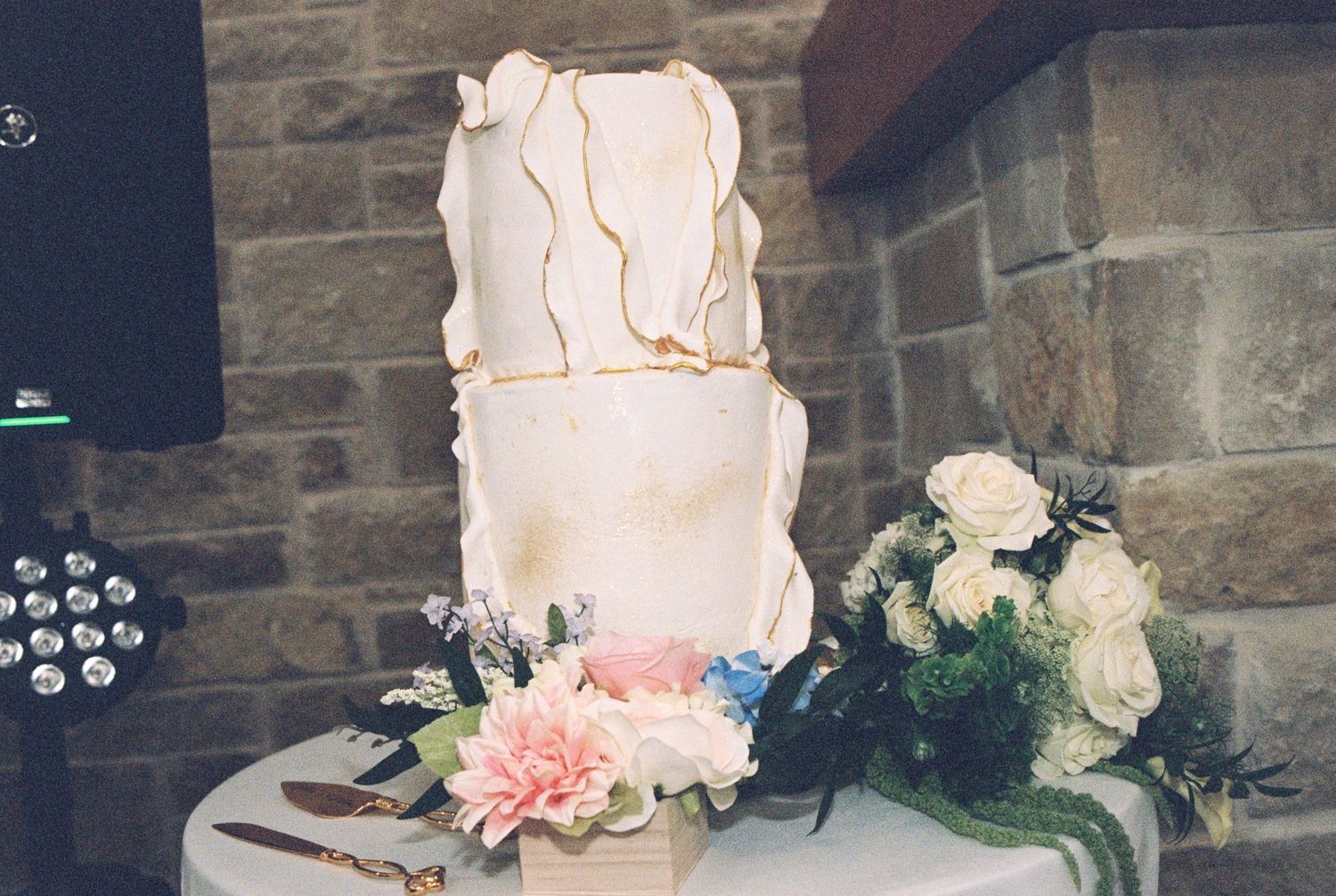An elegant white textured wedding cake decorated with pink and white flowers against a stone wall.