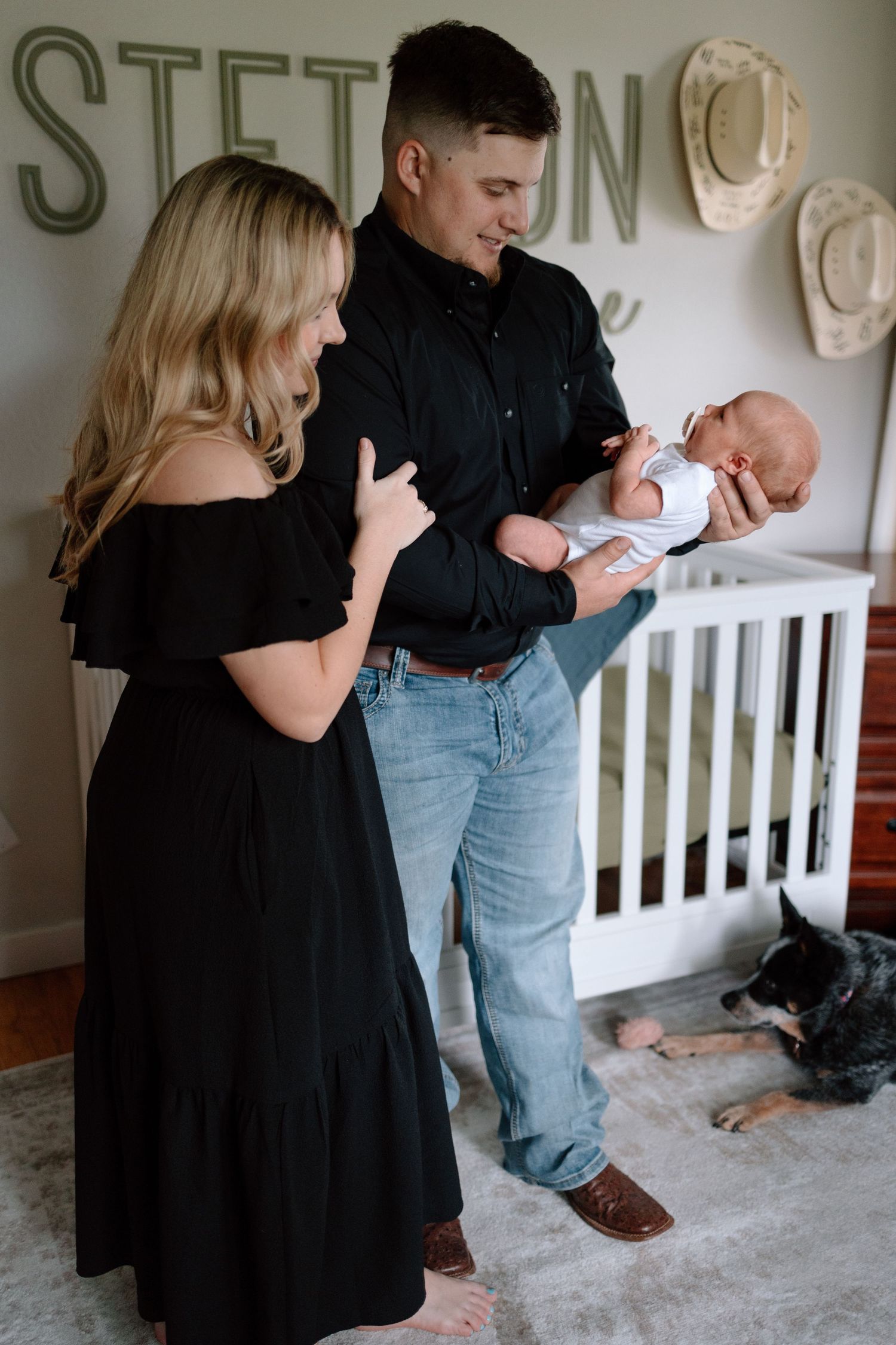 A couple stands in their nursery with a newborn baby while a dog rests on the floor near a white crib.