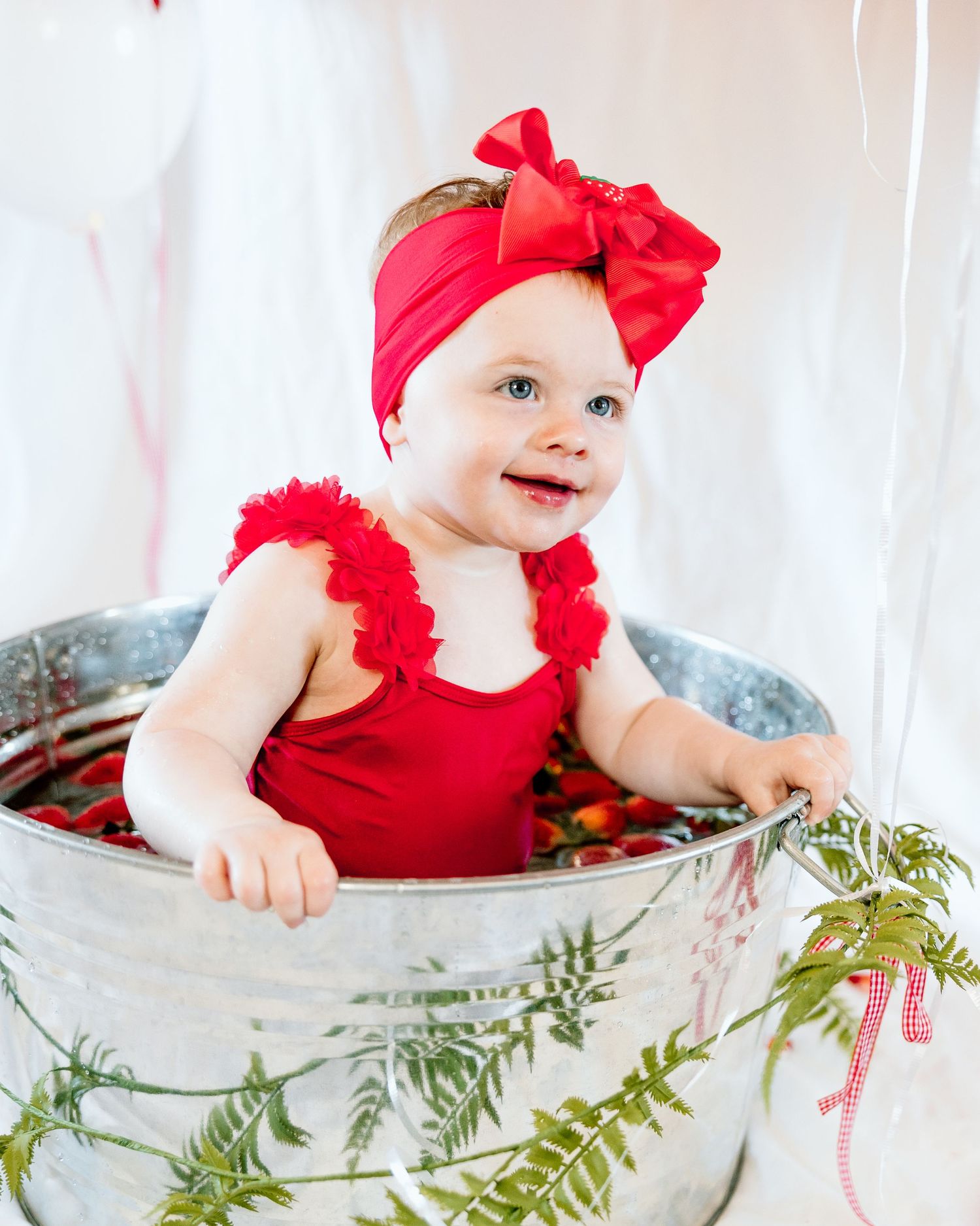 Baby girl in red romper and bow headband sitting in galvanized tub with berries and greenery.