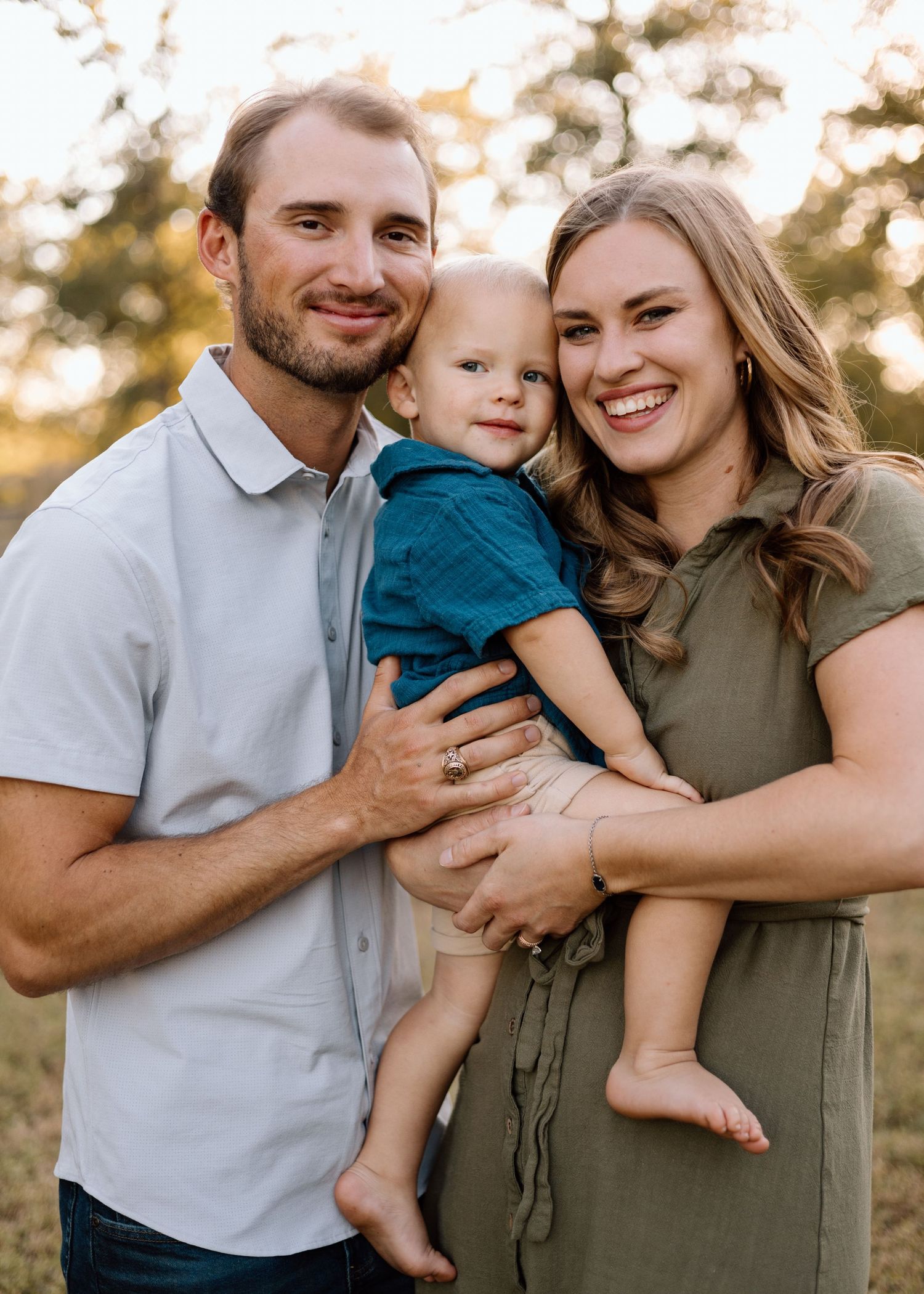 A smiling young family with a man, woman, and baby boy posing outdoors in warm golden light.
