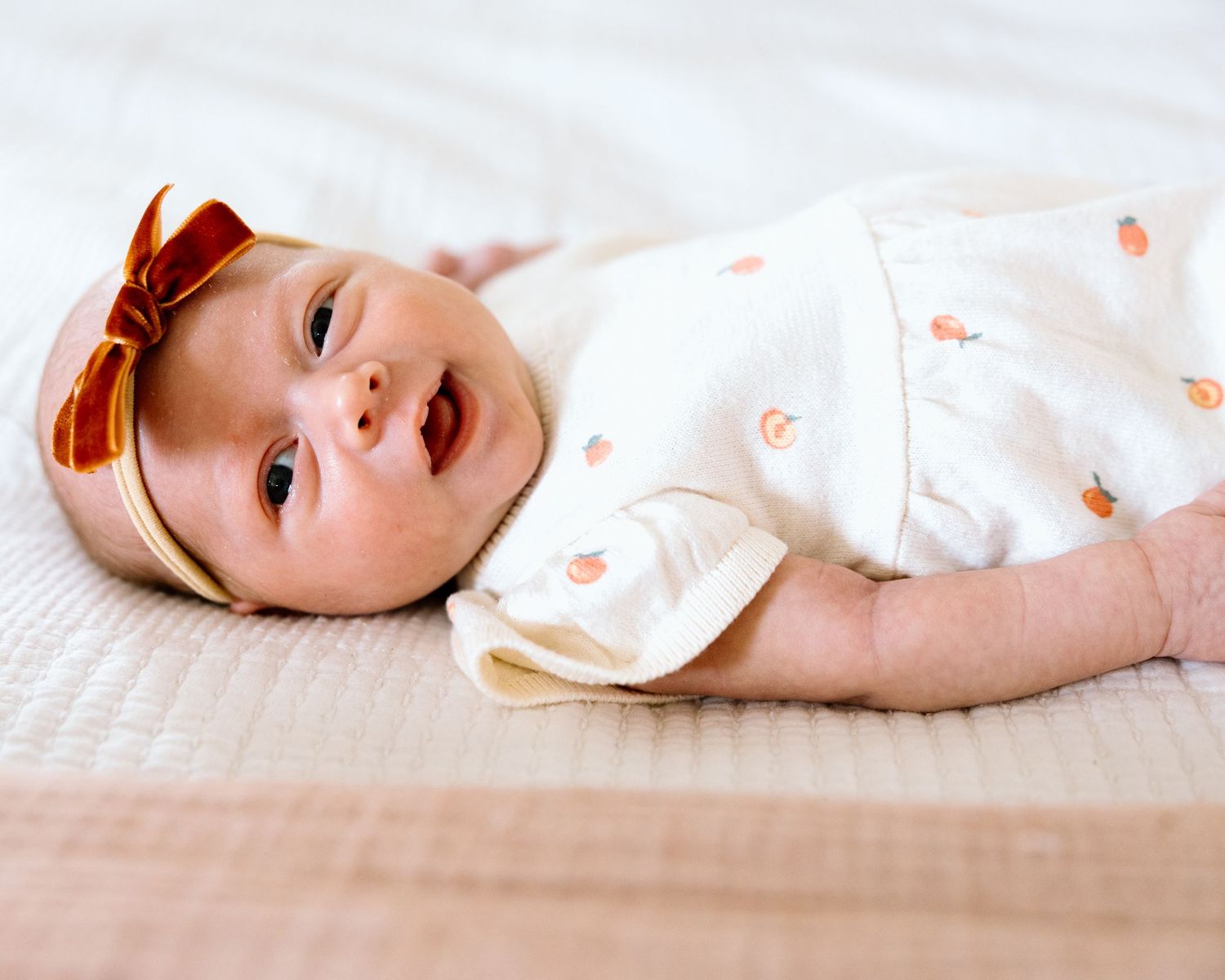 Adorable baby girl in white peach-print outfit and red bow headband lying on a quilted white surface.