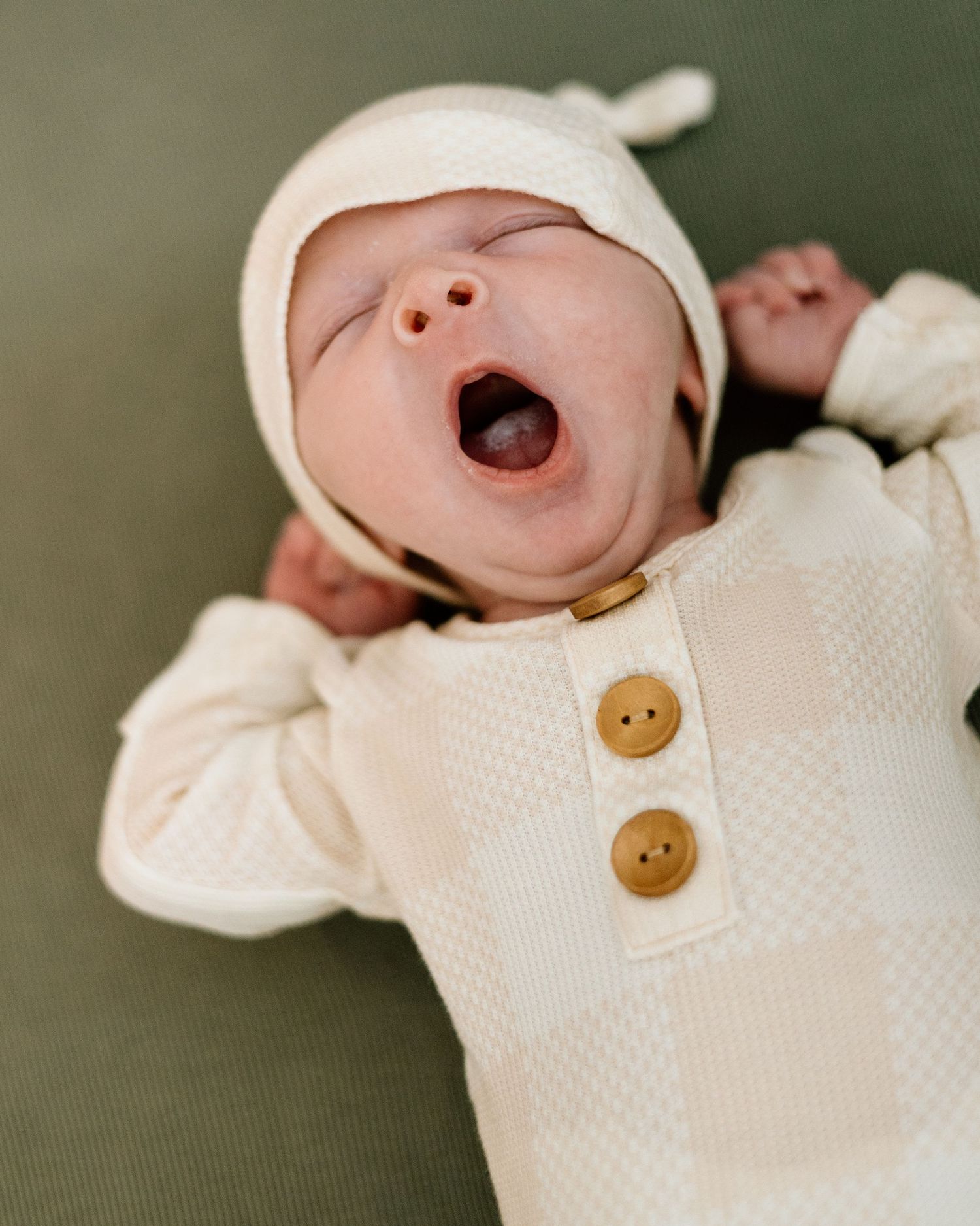 Cute newborn baby yawning in cream knit outfit and bonnet with brown buttons lying on green surface.