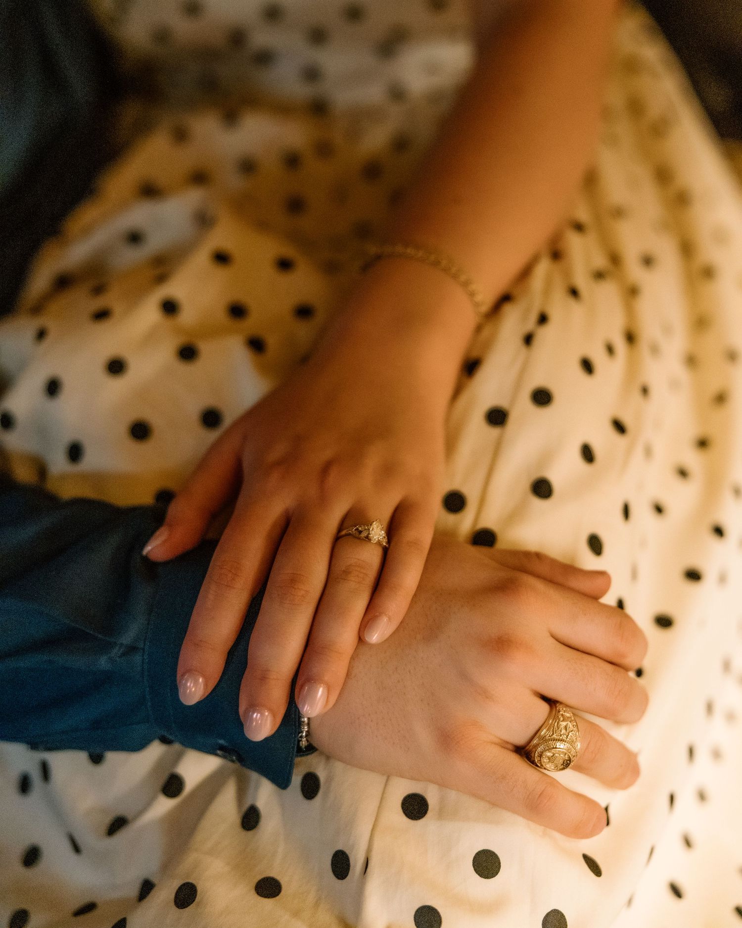 Two hands adorned with gold rings rest on a white polka dot dress, with a delicate gold bracelet visible.