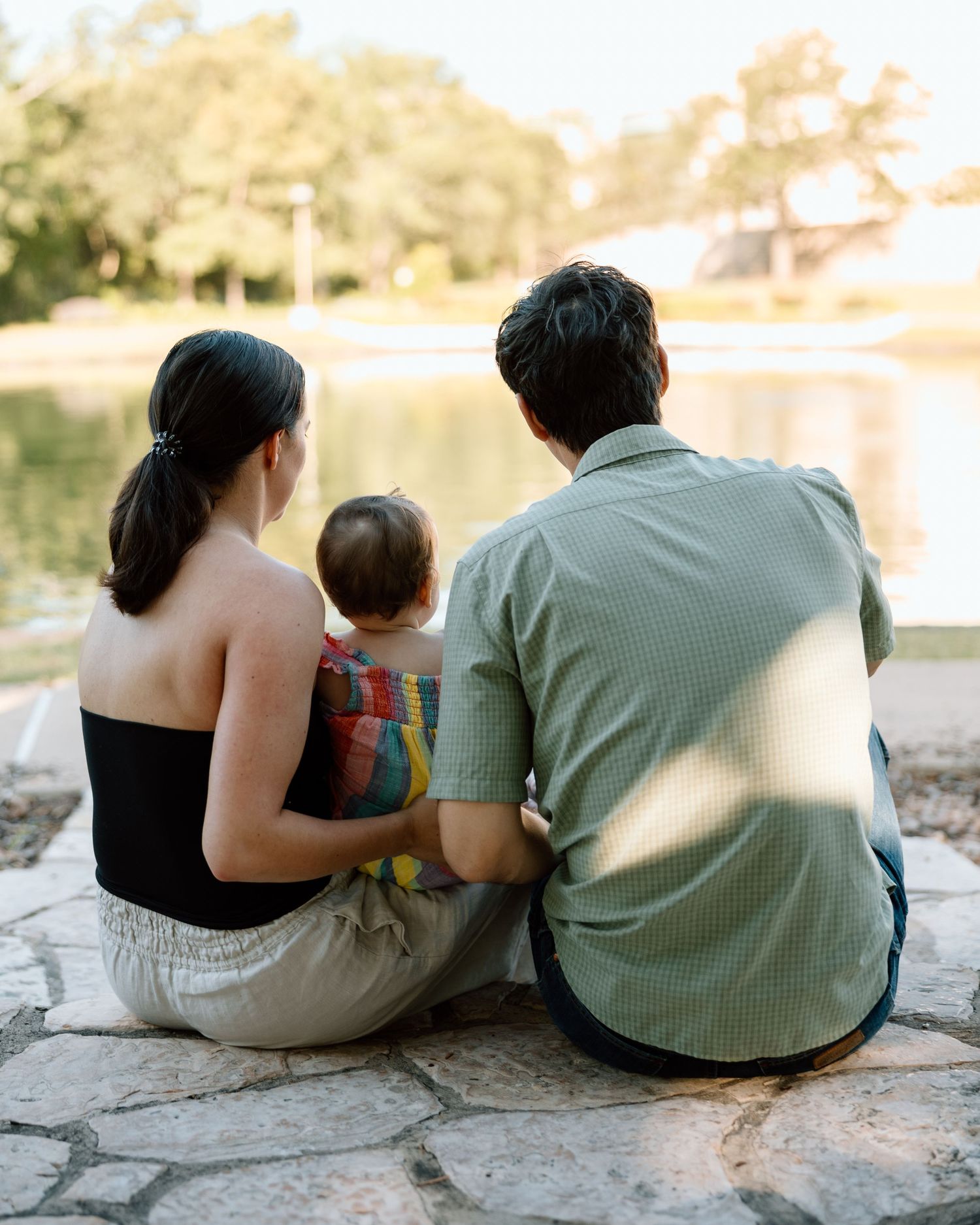 Family of three sitting on stone pavement by a peaceful lake, viewed from behind on a sunny day.