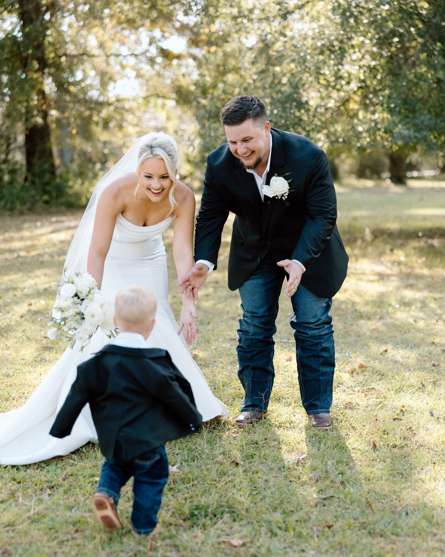 Laughing bride and groom in outdoor setting greet a small child in a tiny black suit.