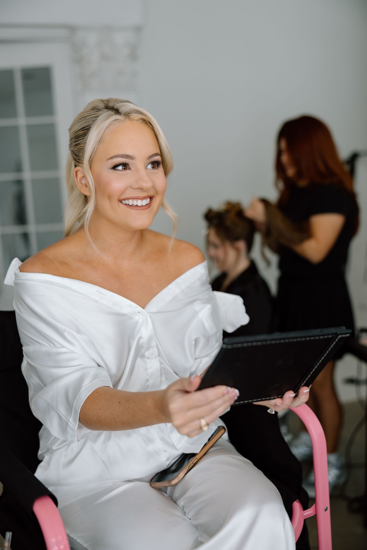Smiling blonde bride in white off-shoulder robe holds makeup palette while getting ready in a bright bridal suite.