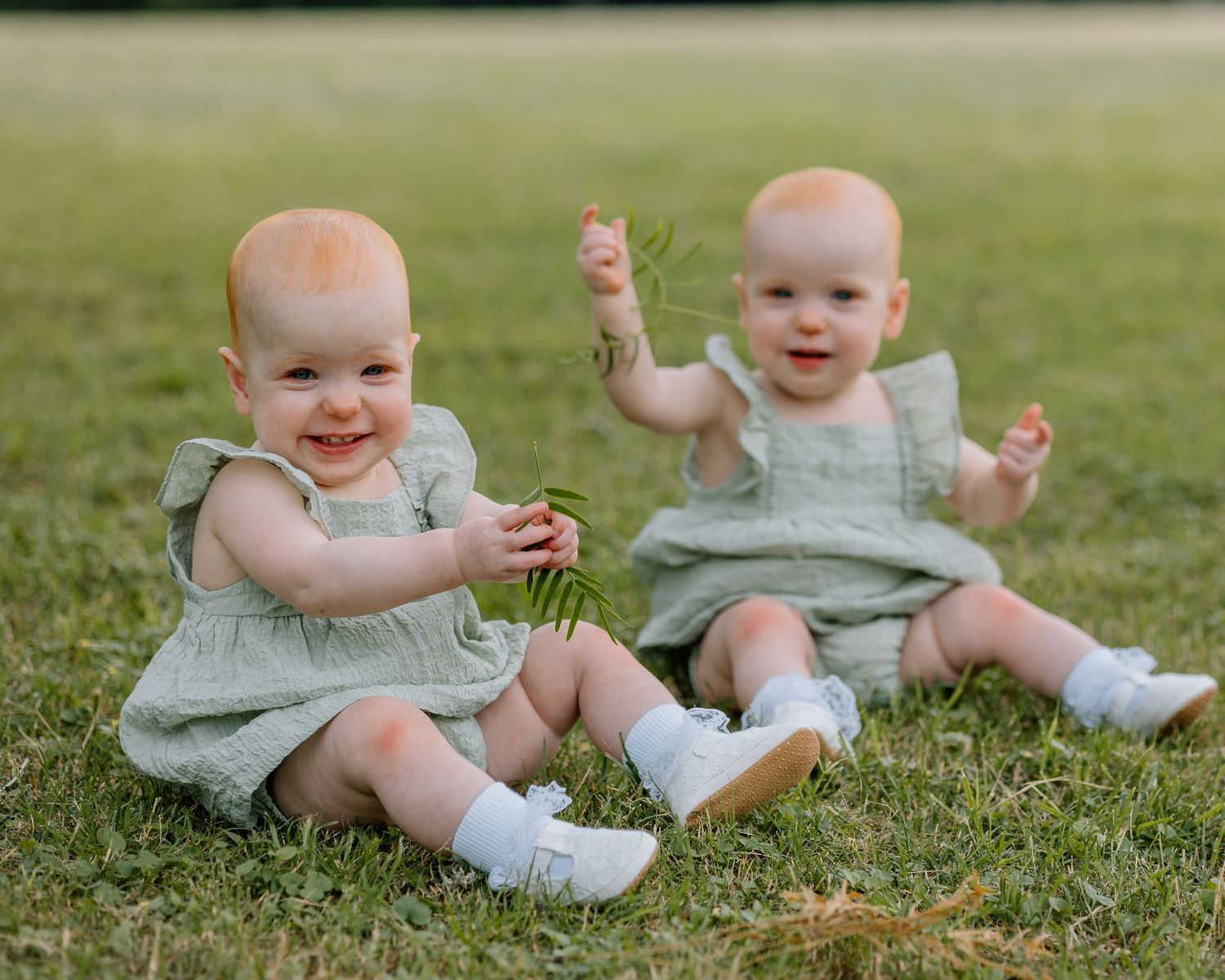 Two babies in matching mint green dresses sit on grass smiling and playing together outdoors.