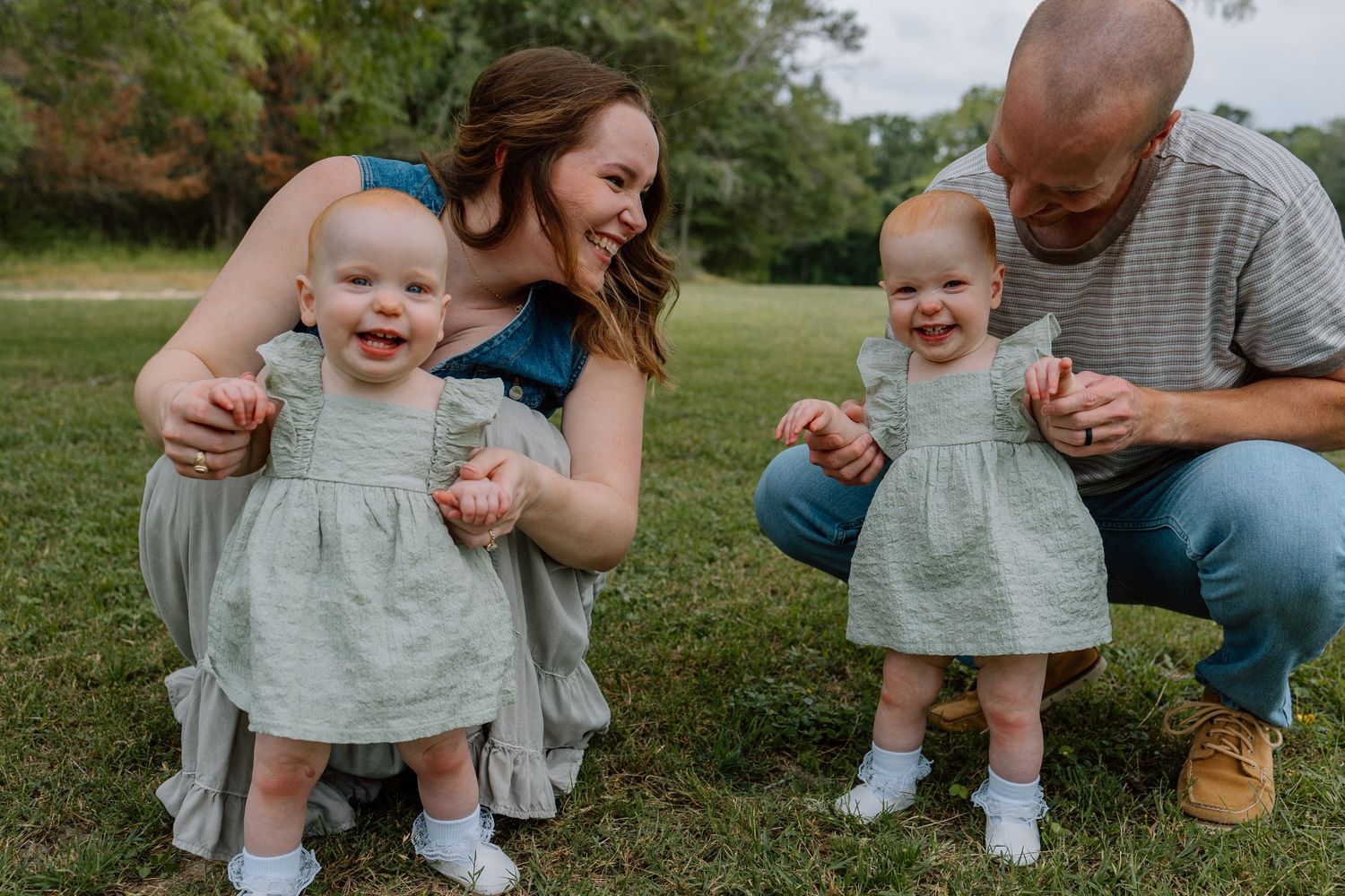 Outdoors on grass, a family group shares a joyful moment with twin toddlers wearing matching gray dresses.