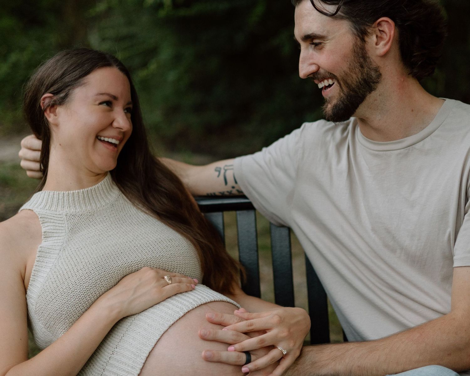 Expectant couple sharing joyful laughter while sitting on a wooden bench outdoors.