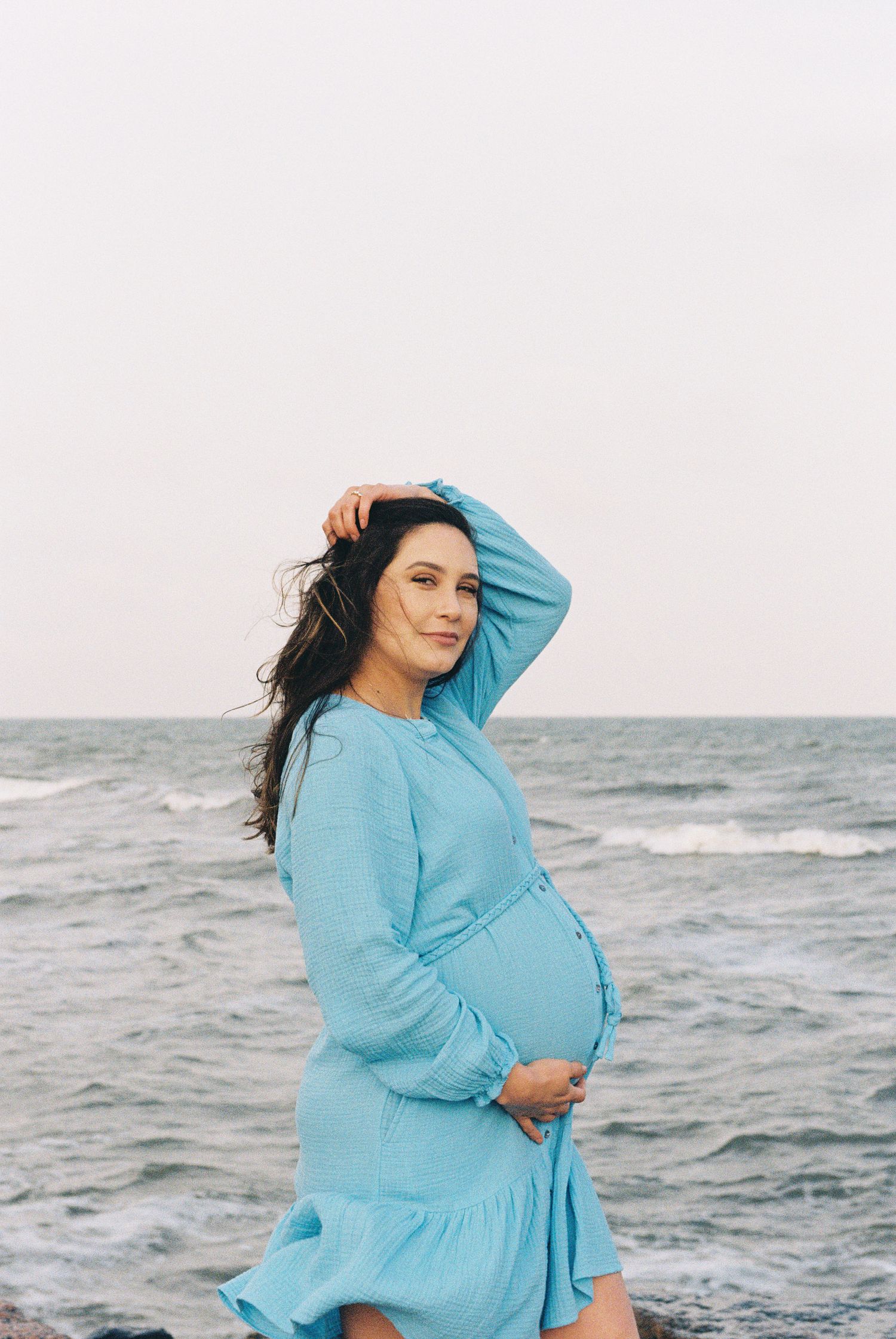 A pregnant woman in a flowing blue dress poses against an ocean backdrop.