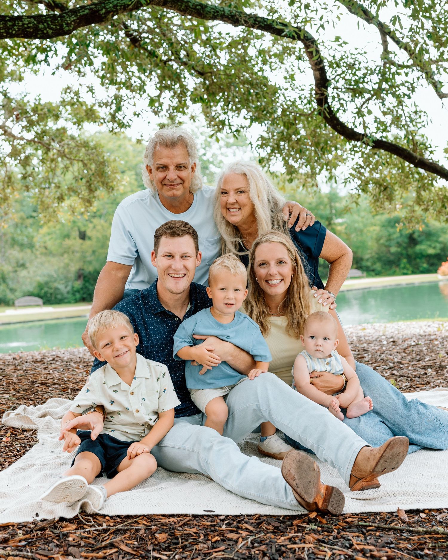 Multi-generational family portrait with grandparents, parents, and three young children sitting outdoors near a pond.