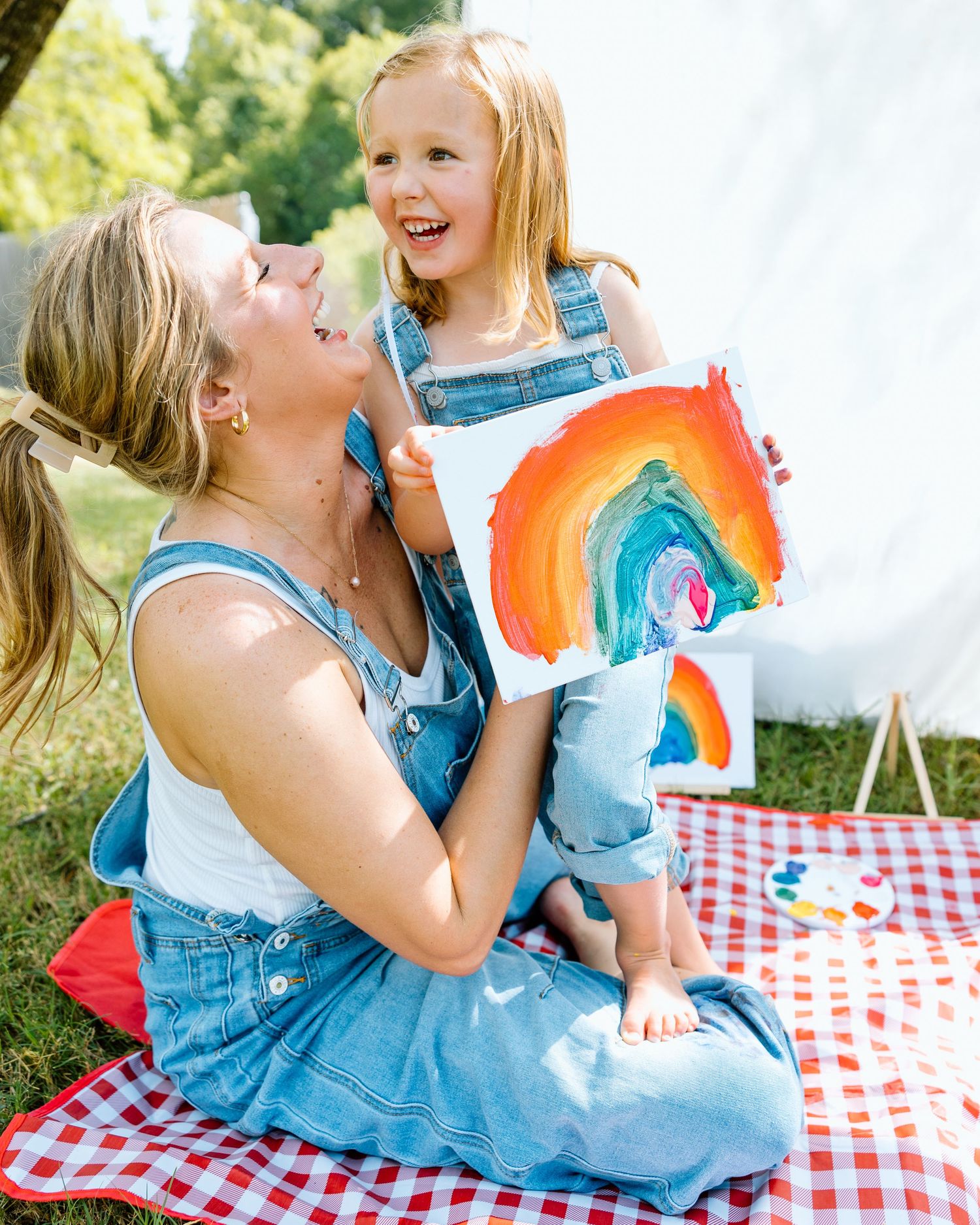 Proud little girl shows colorful rainbow painting to smiling mom on red gingham picnic blanket outdoors.