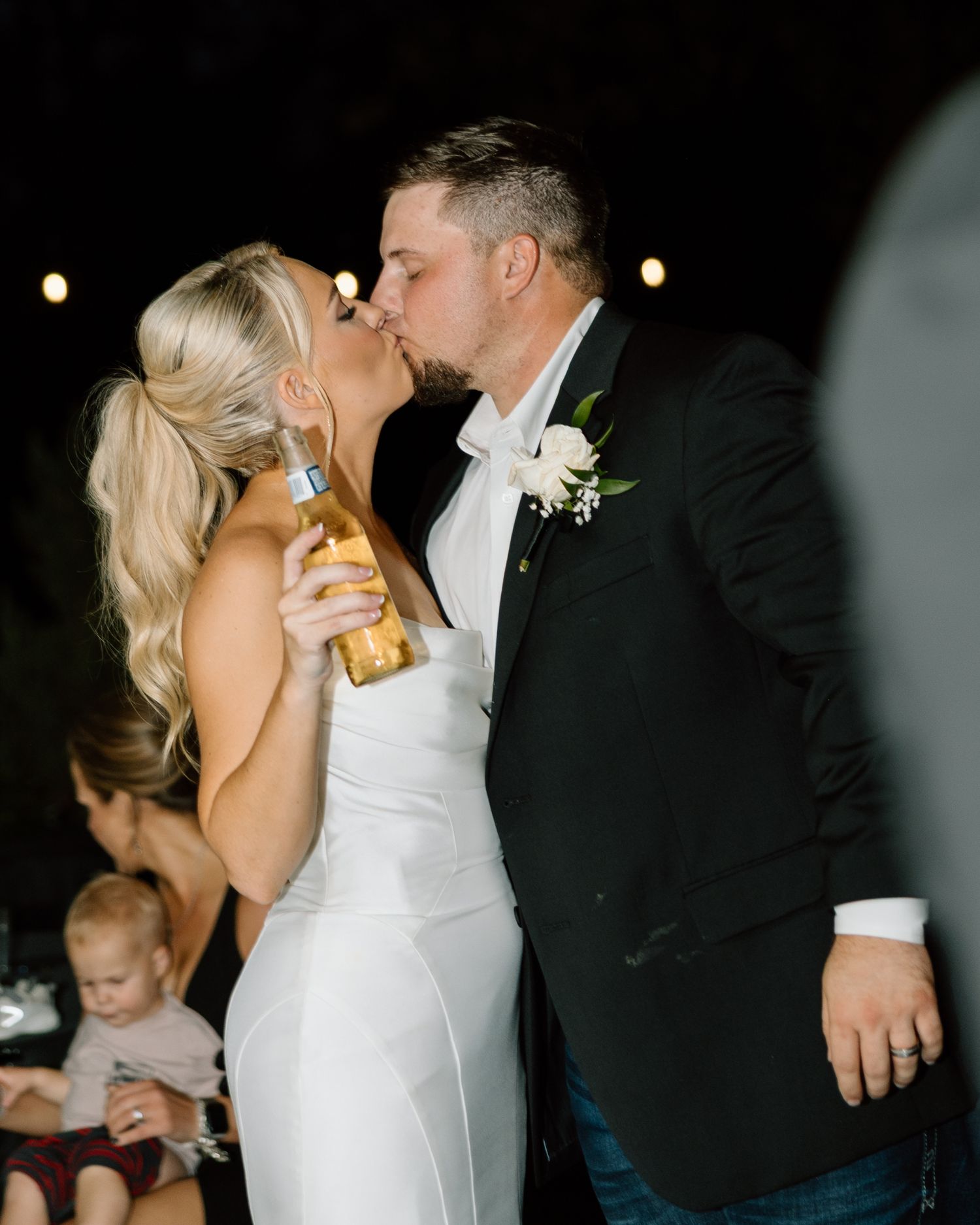 Bride and groom share a kiss at their wedding reception, bride holding a beer bottle, groom in black tuxedo with boutonniere.
