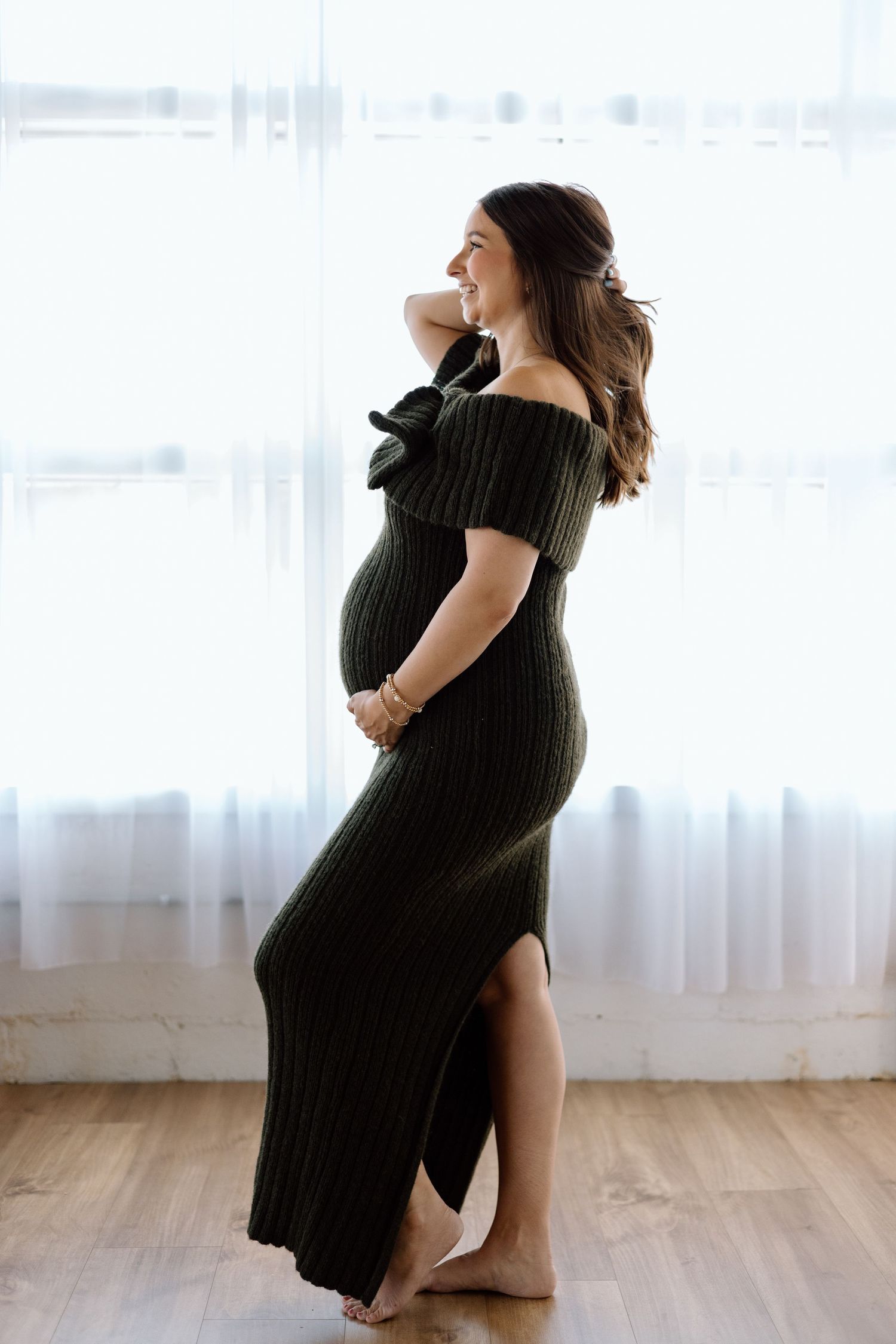 Pregnant woman in olive green off-shoulder ribbed maxi dress standing by window, cradling baby bump.